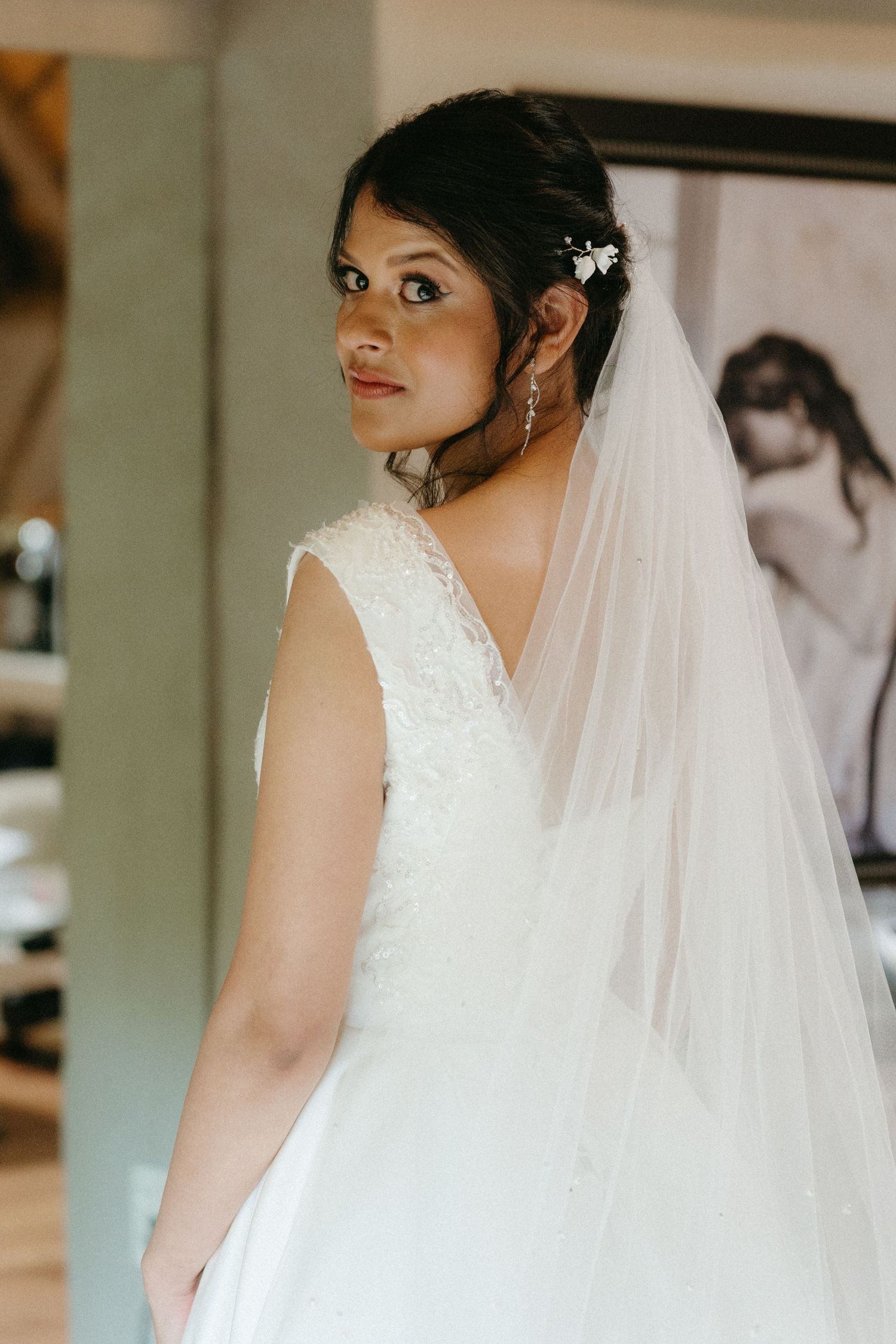 Bride wearing elegant lace wedding dress and long flowing veil looks over shoulder in soft natural light.