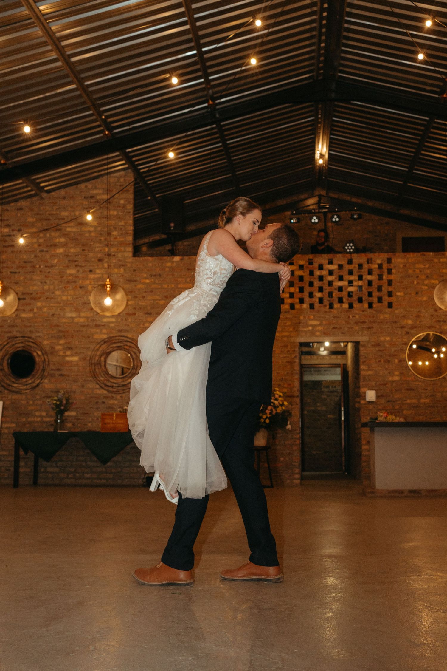 Newlyweds share a dynamic dancing moment under industrial pendant lights in a brick venue.