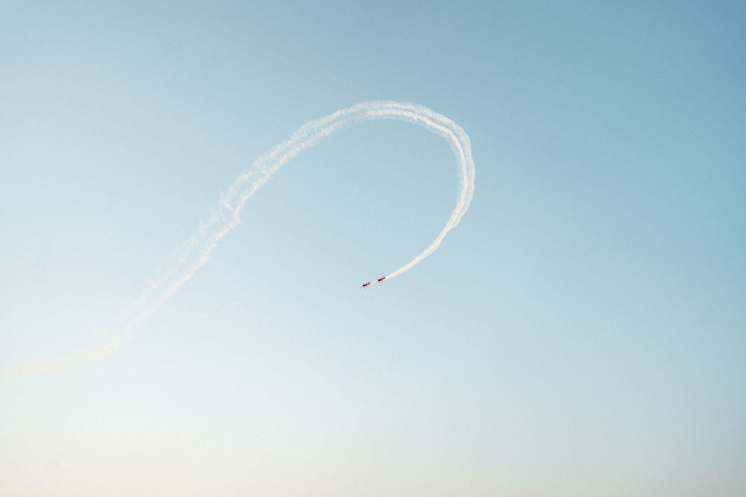 An airplane creates a graceful arc of vapor trails against a soft blue sky.