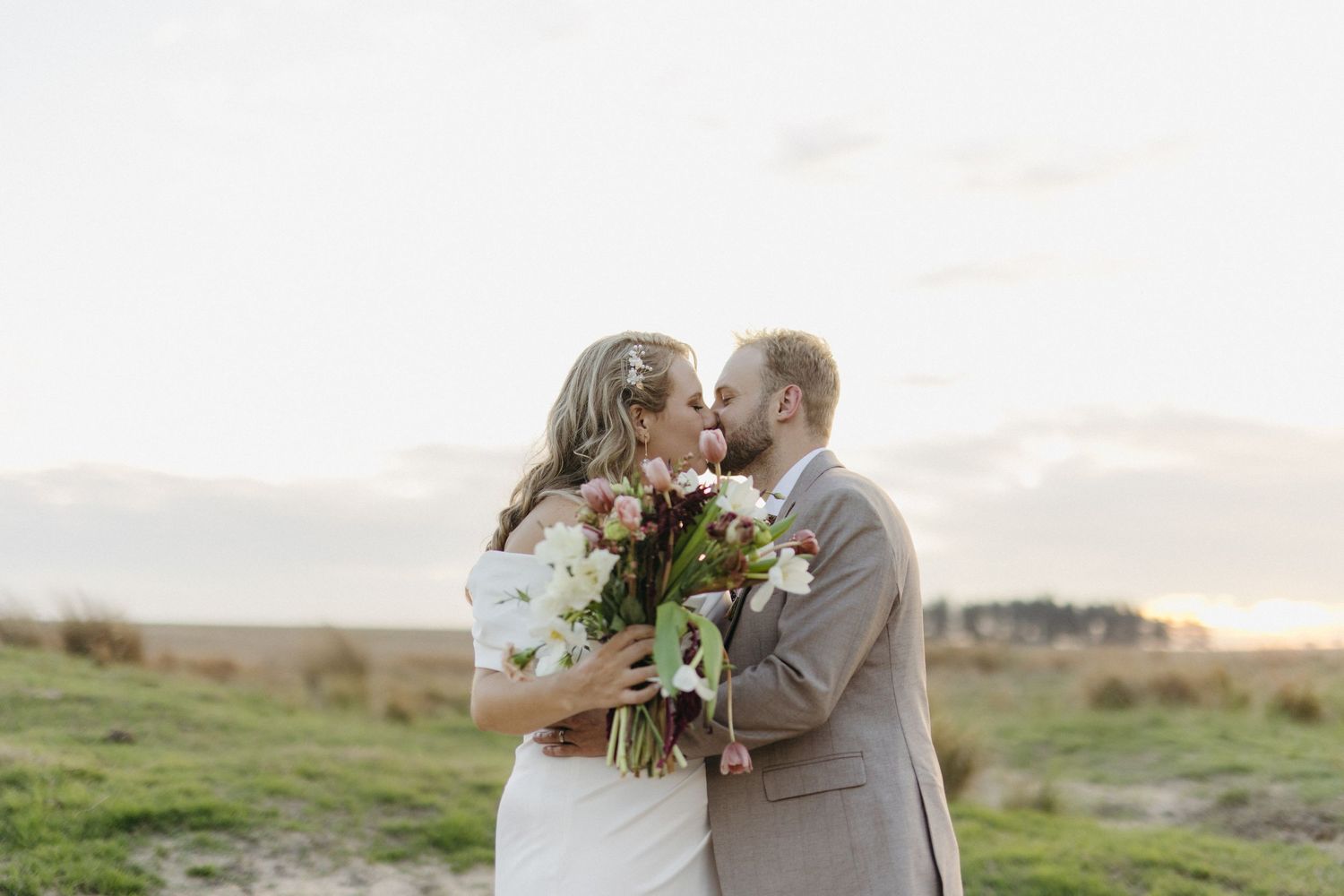 A couple shares a romantic moment while holding a beautiful bouquet in an outdoor field at sunset.