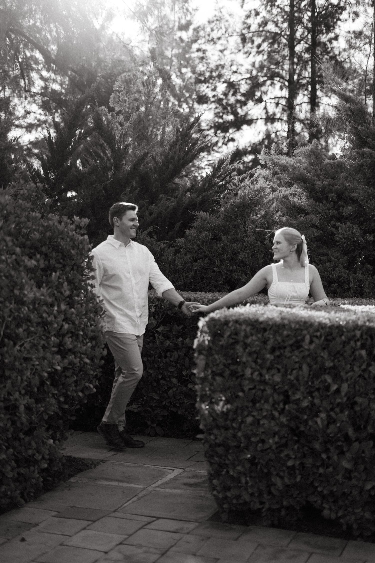Romantic couple holding hands while walking through garden pathway with manicured hedges.