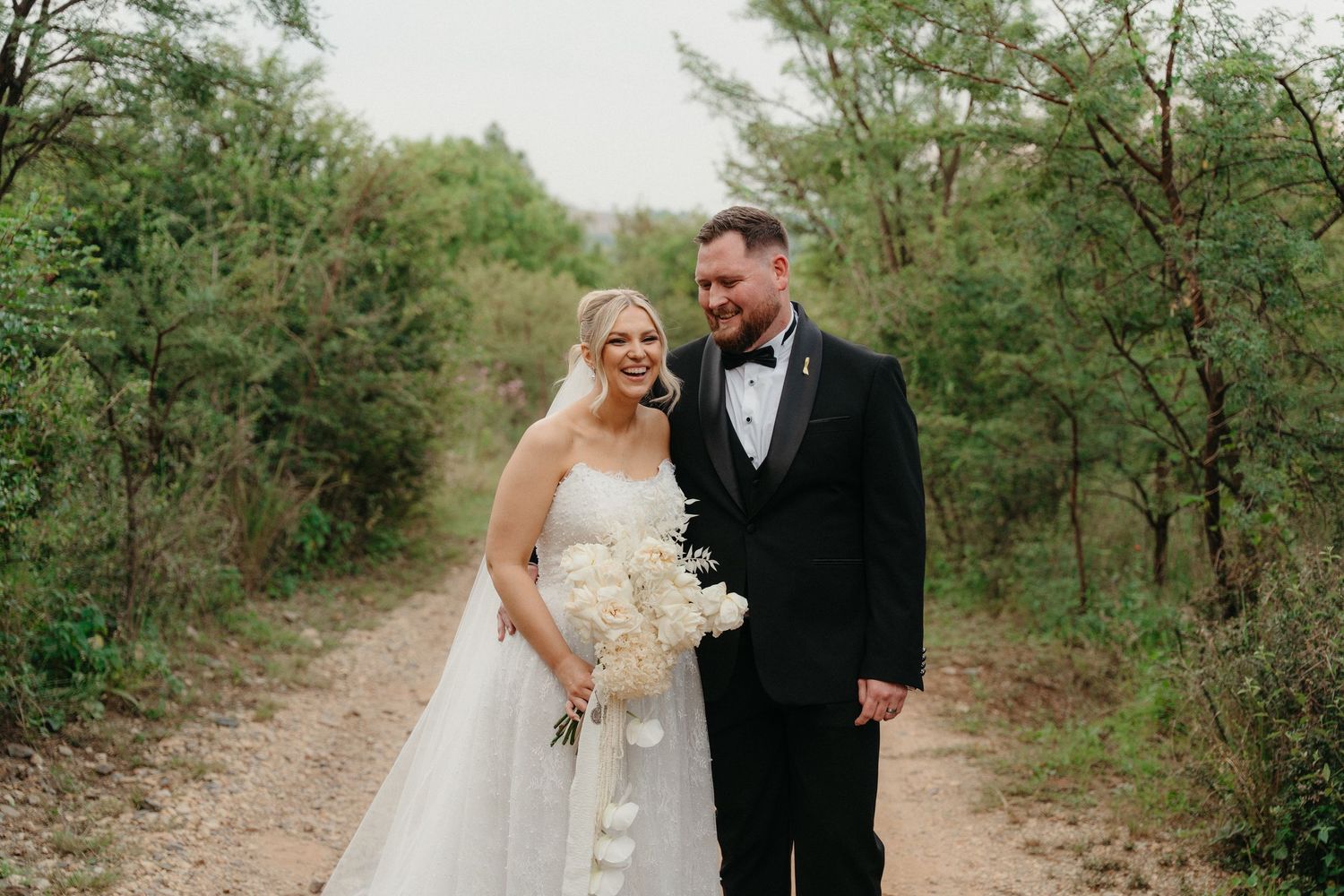A couple in wedding attire shares romantic moments along a tree-lined outdoor path during their special day.