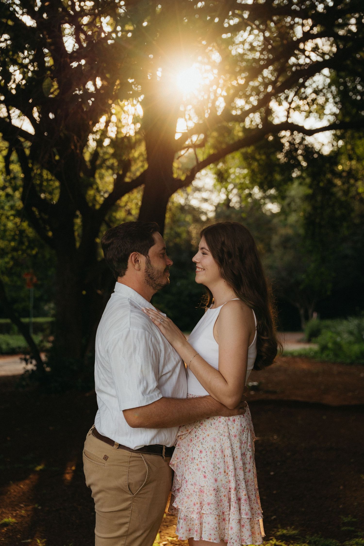 Couple embracing at golden hour in a lush park, warm sunlight glowing through trees behind them.