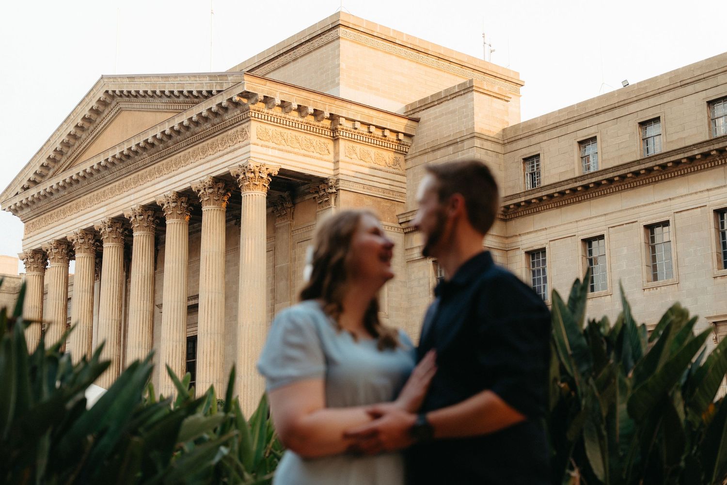 A couple shares a romantic moment in front of a grand neoclassical building with columns at sunset.