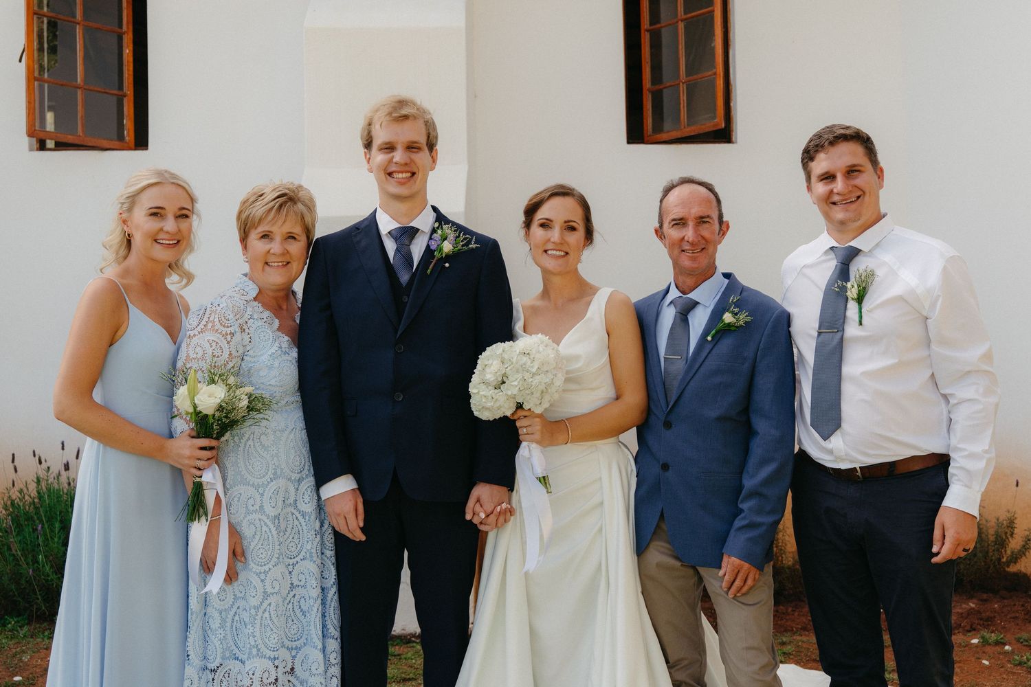 Wedding party members pose together in formal attire outside a white building with brown window frames.