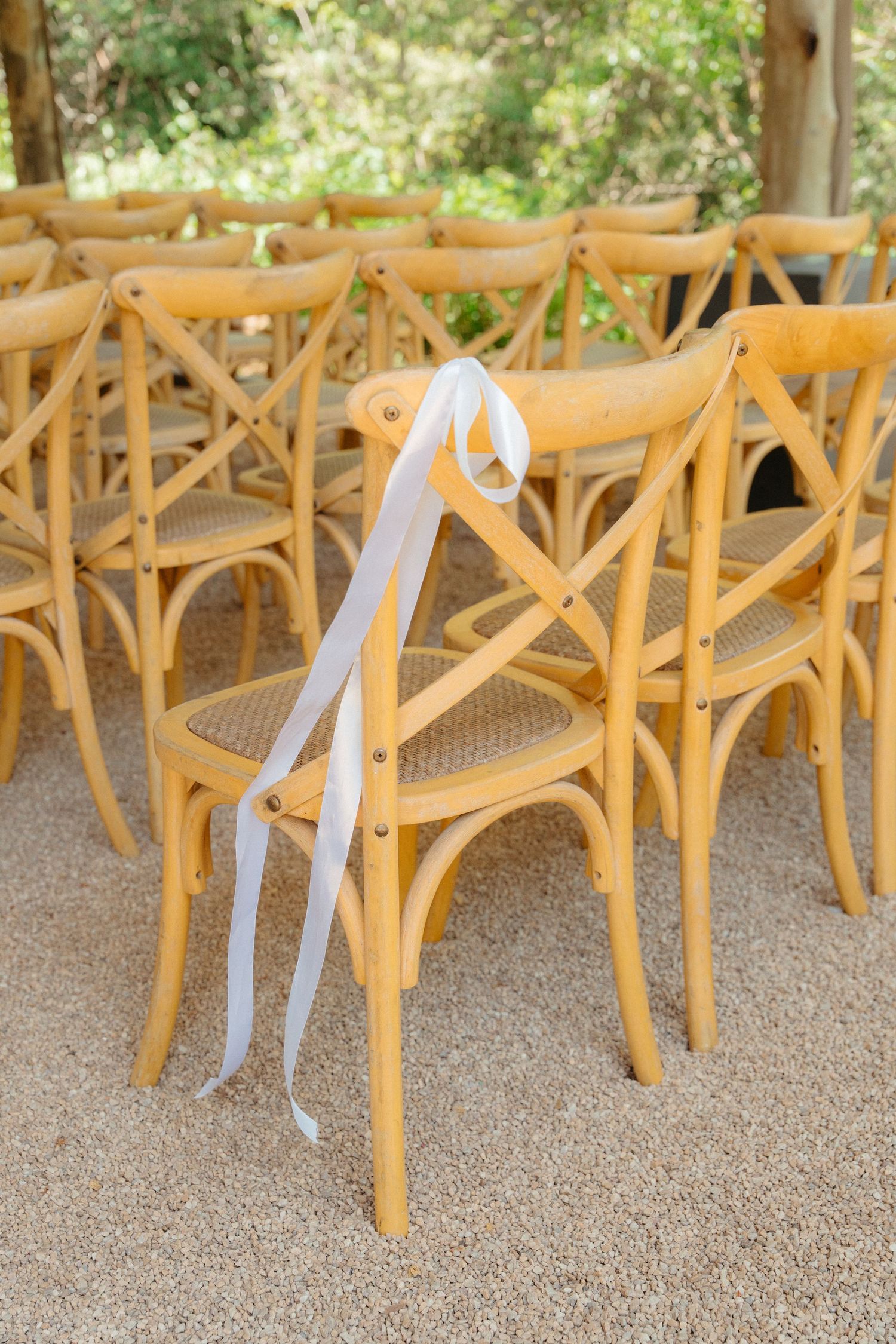 Wooden cross-back chairs with white ribbons arranged in rows for an outdoor wedding ceremony.
