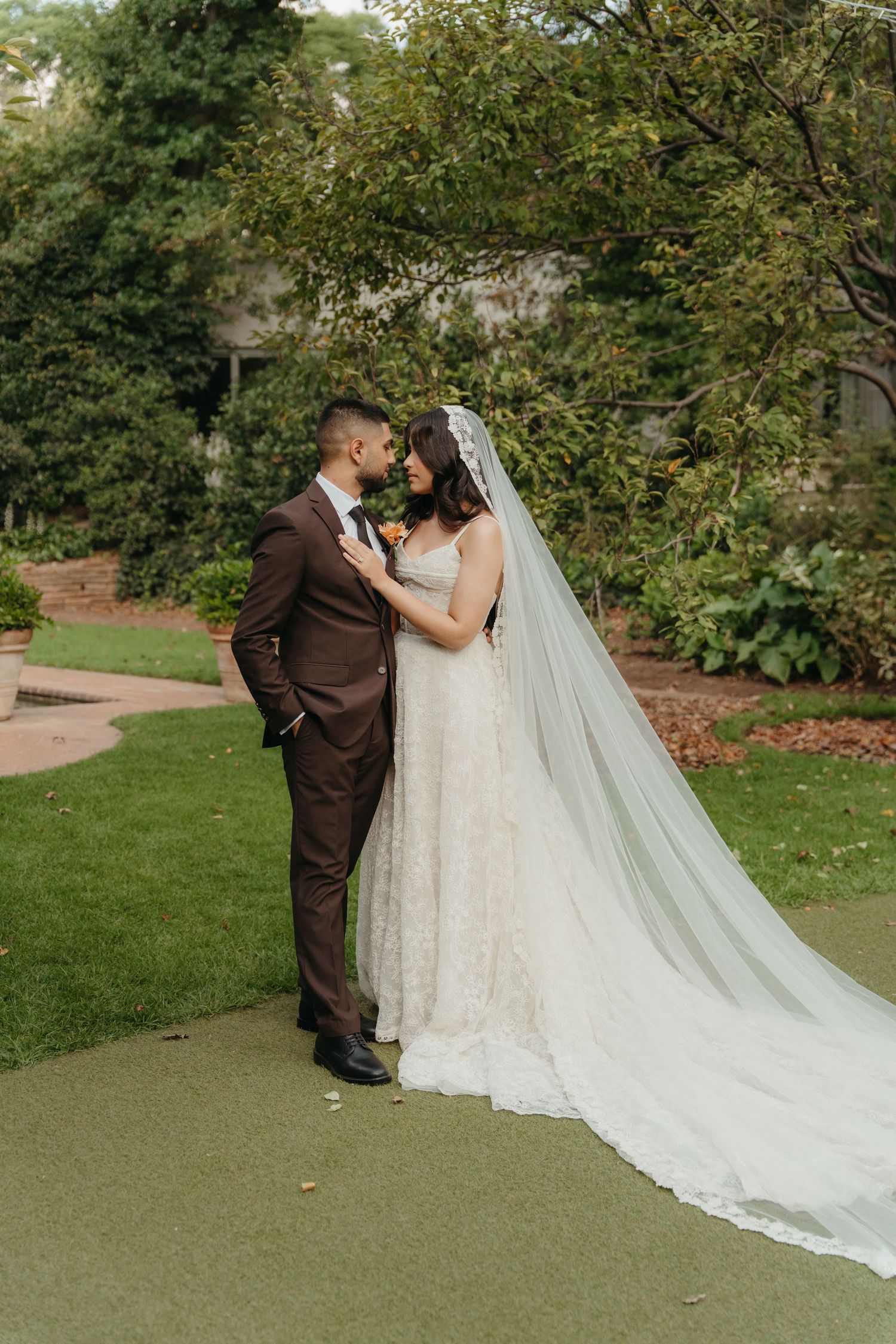 A bride in a lace gown and long veil stands with a groom in a brown suit in a lush garden setting.