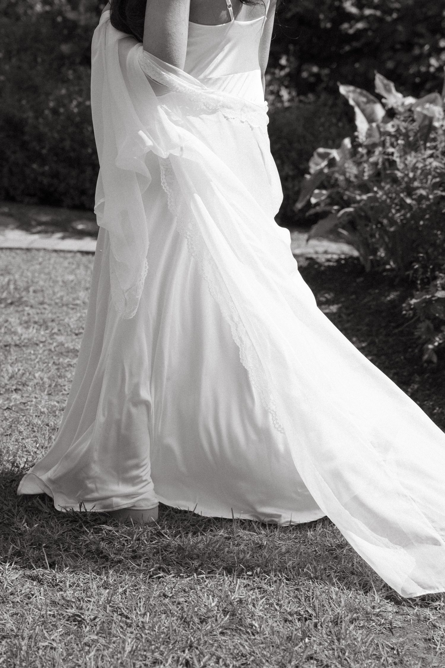 Black and white photo of a bride in a flowing white wedding gown standing on grass in a garden.