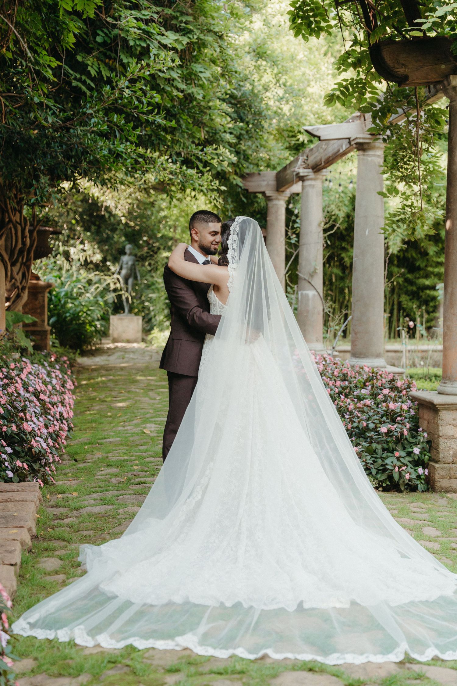 Bride and groom embracing in a lush garden, her long lace veil trailing on stone path near elegant columns.
