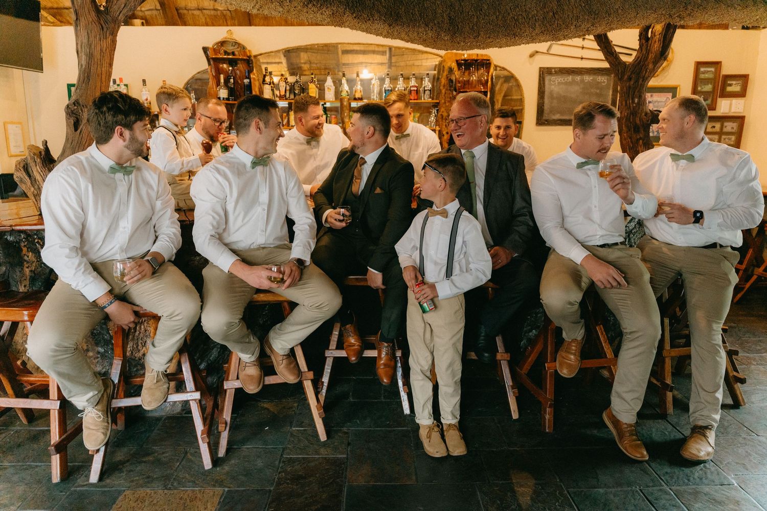 Group of groomsmen in matching white shirts and gray pants sitting on wooden stools at a reception.