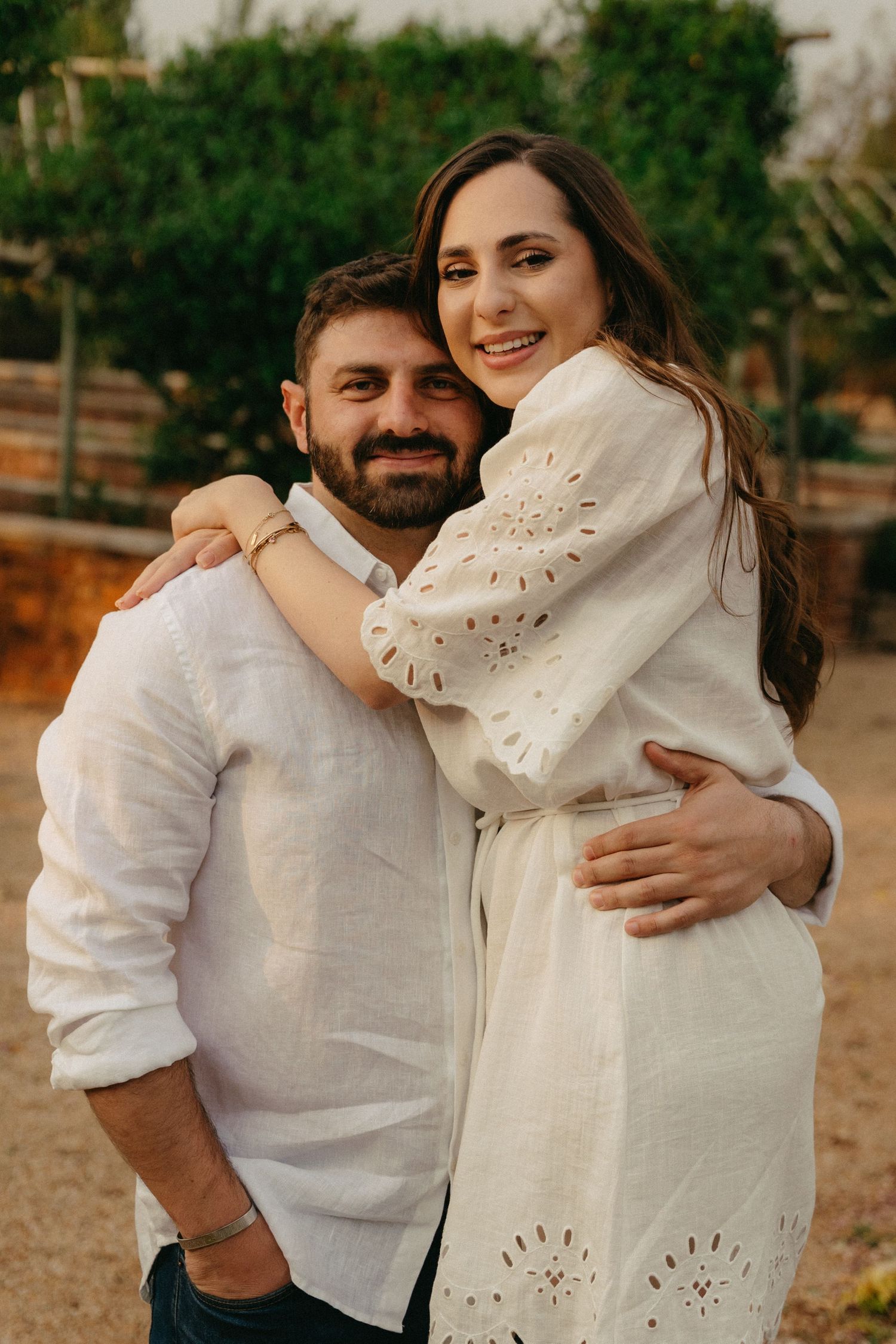 Romantic couple in white clothing embracing outdoors against a natural green background.