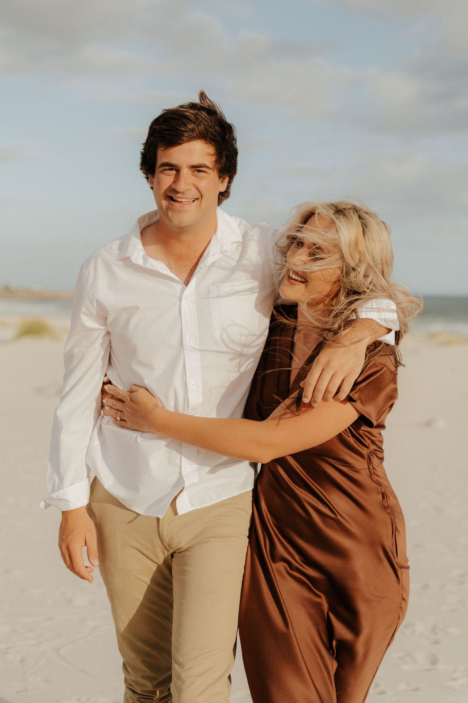 Happy couple walking on beach, man in white shirt and khakis, woman in brown satin dress, smiling.