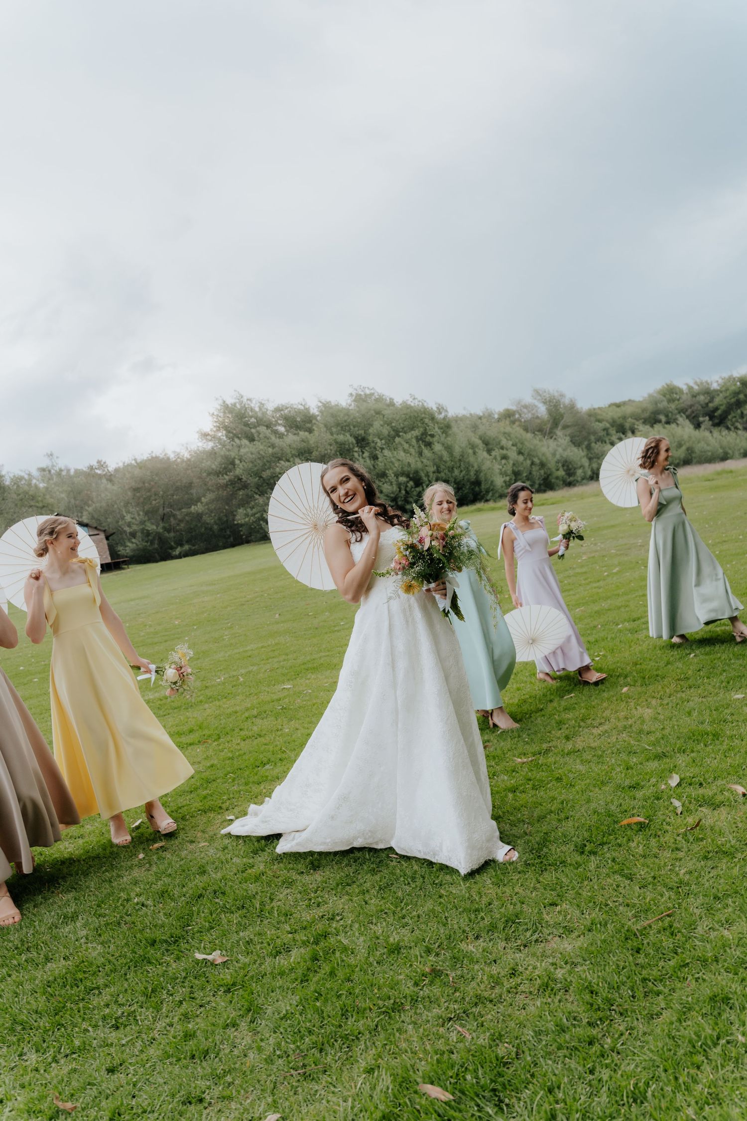 A wedding party in yellow dresses runs playfully across a green hillside field holding white parasols.