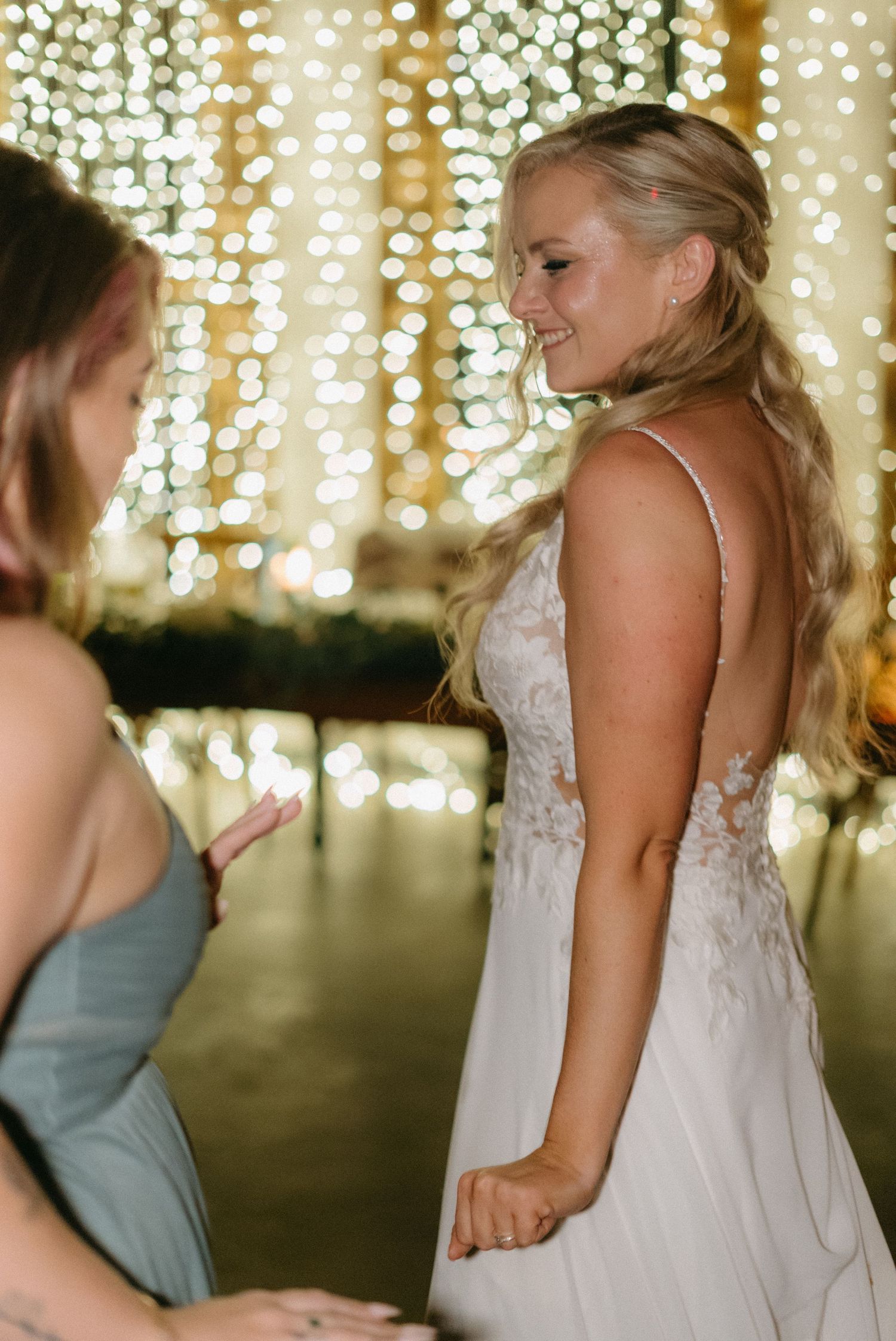 A bride in a white lace dress smiles joyfully against a backdrop of twinkling string lights.