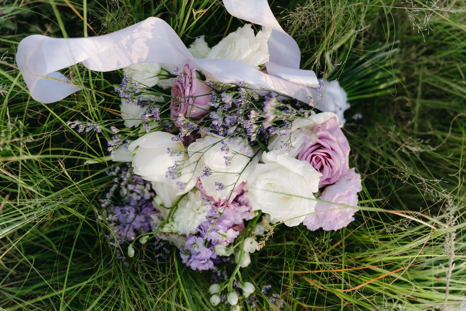 A delicate wedding bouquet of white and lavender flowers resting in green grass with a flowing ribbon.