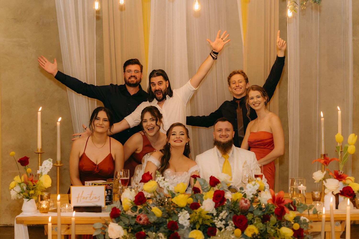 Wedding party poses joyfully behind a decorated reception table with floral centerpieces.
