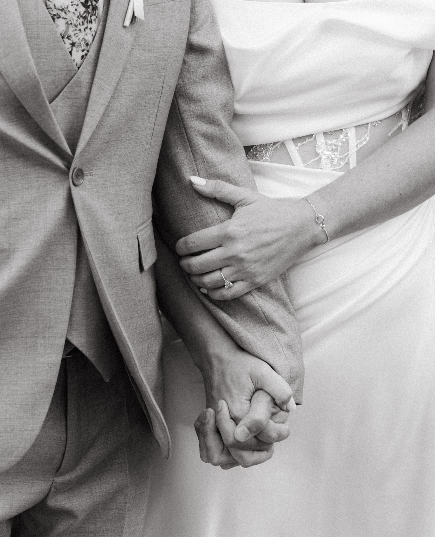 Black and white close-up of a newlywed couple holding hands, showing wedding attire with a gray suit and white dress.