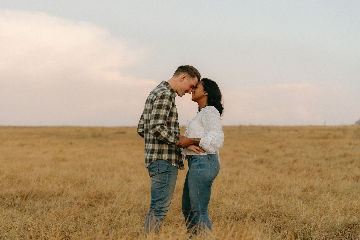A couple stands together in a golden wheat field at sunset creating a romantic silhouette against the warm sky.