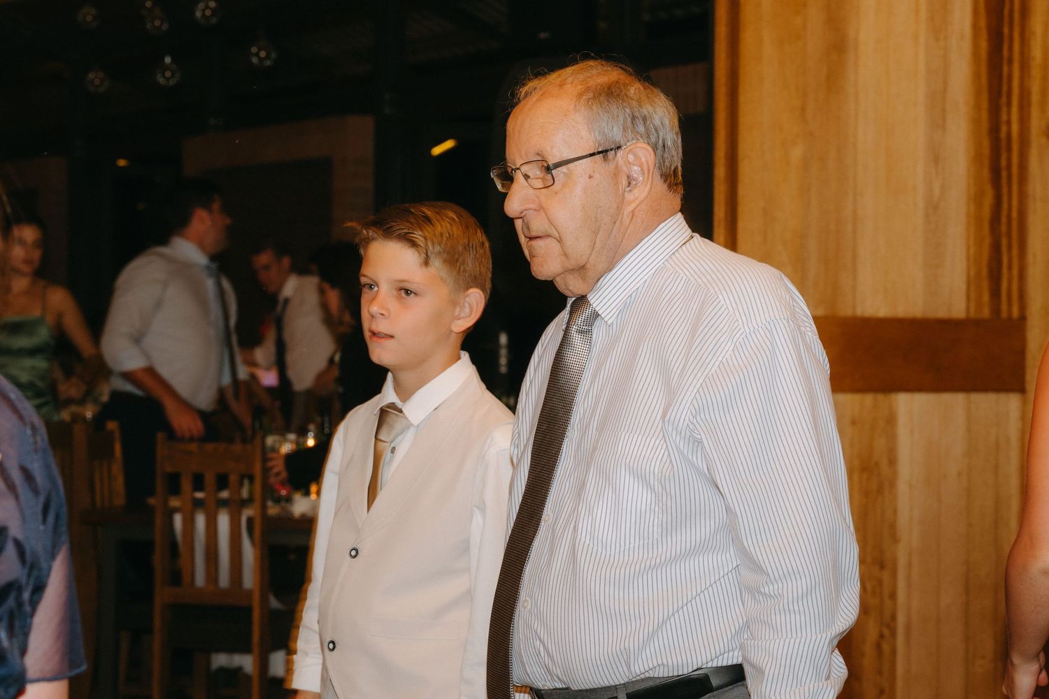 Young boy in white dress shirt and older gentleman in white dress shirt and tie standing together at formal event.