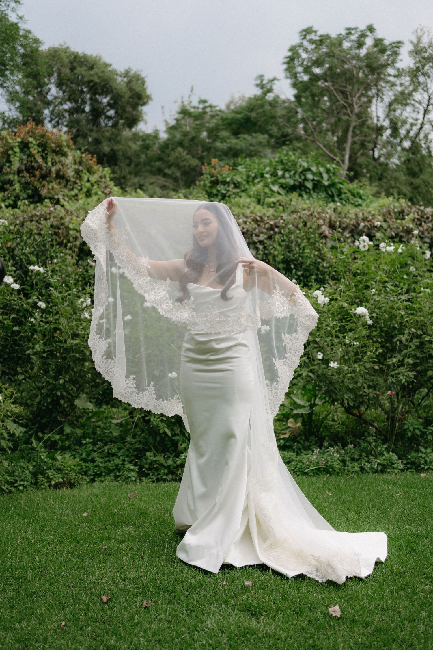 A bride in a fitted white wedding gown poses gracefully with her flowing veil in a garden setting.