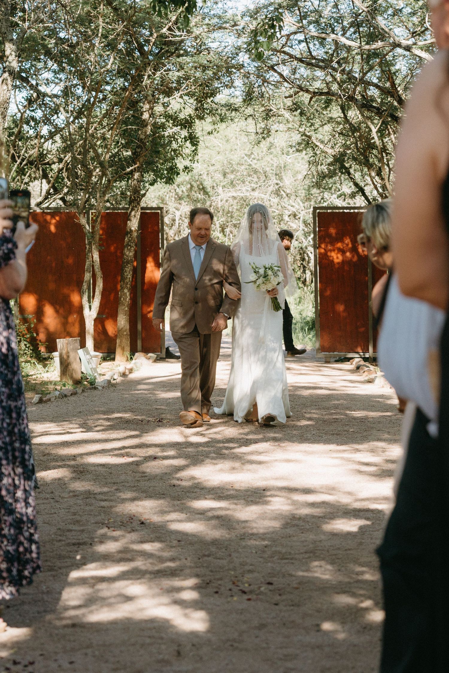 Wedding ceremony processional under dappled sunlight and trees with red doors in background.