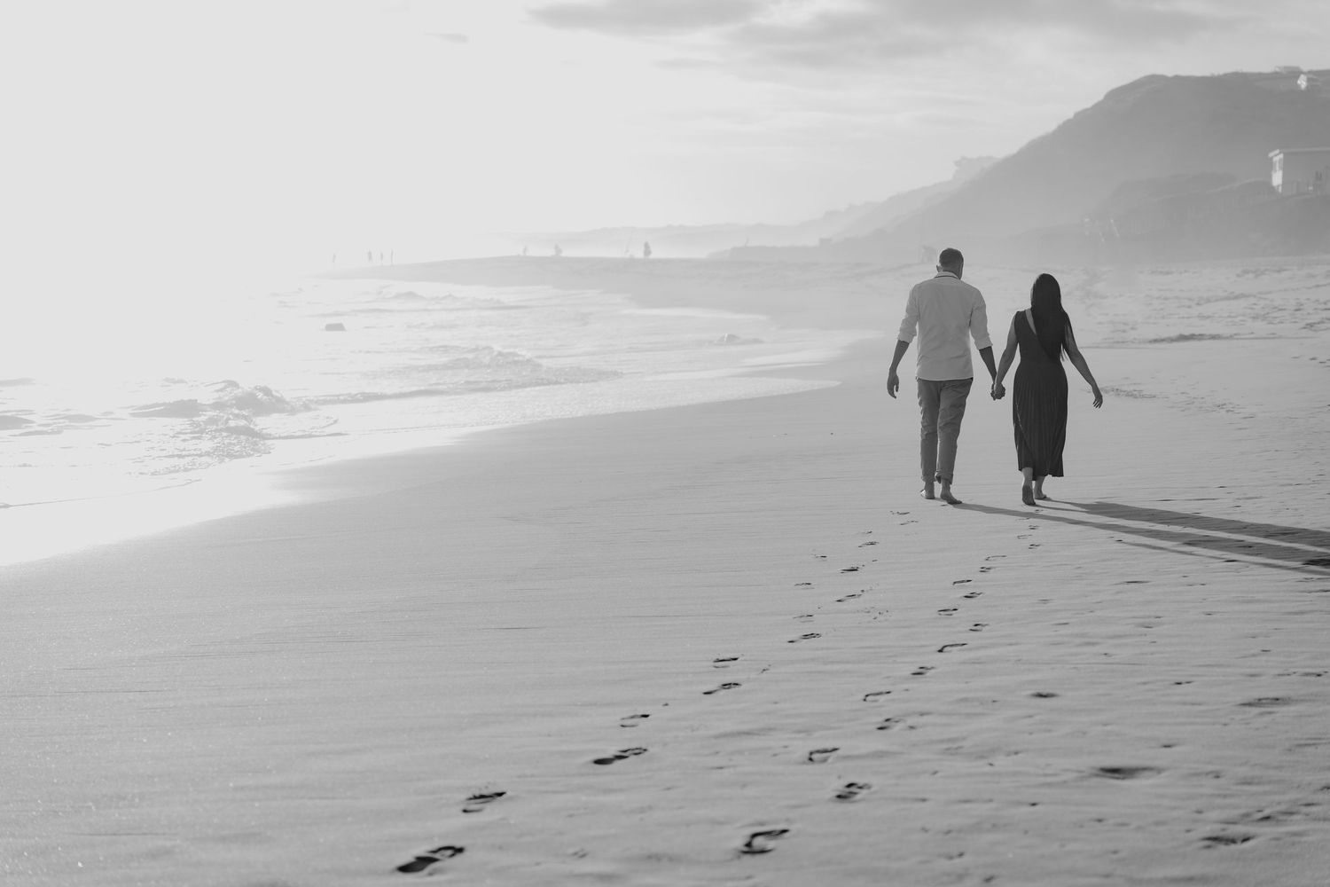 Two silhouettes walk hand in hand along a misty beach at sunset in Hartenbos, Western Cape leaving footprints in the sand.