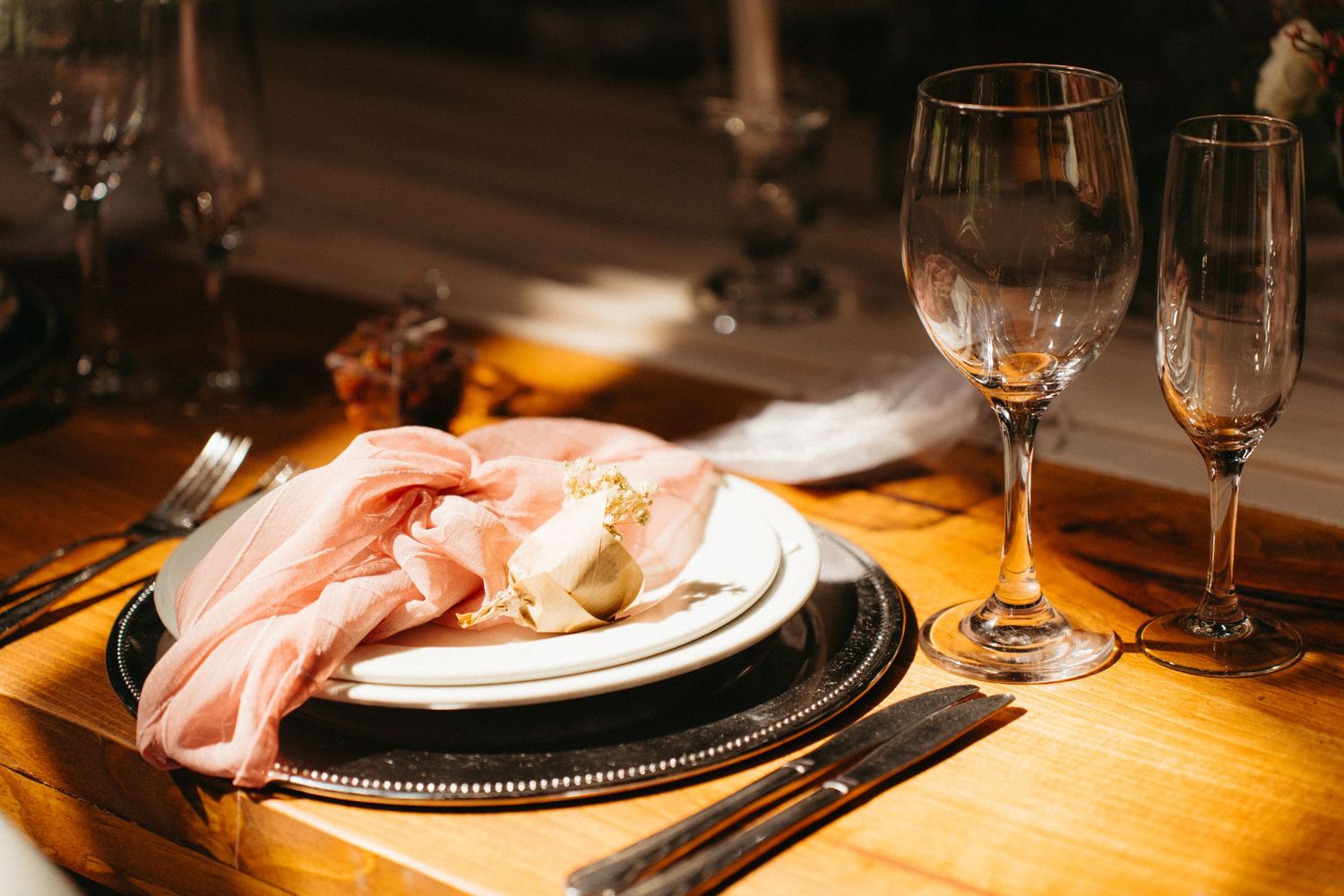 An elegant table setting with wine glasses, candles, and a pink cloth napkin on a wooden surface in warm lighting.