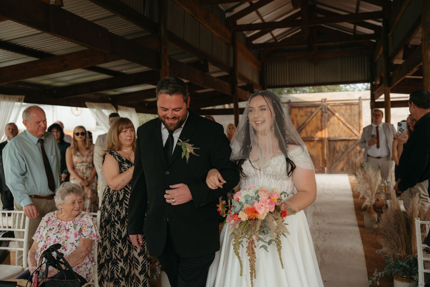 A bride walks down an aisle in a rustic barn venue accompanied by a person while guests watch from the sides.