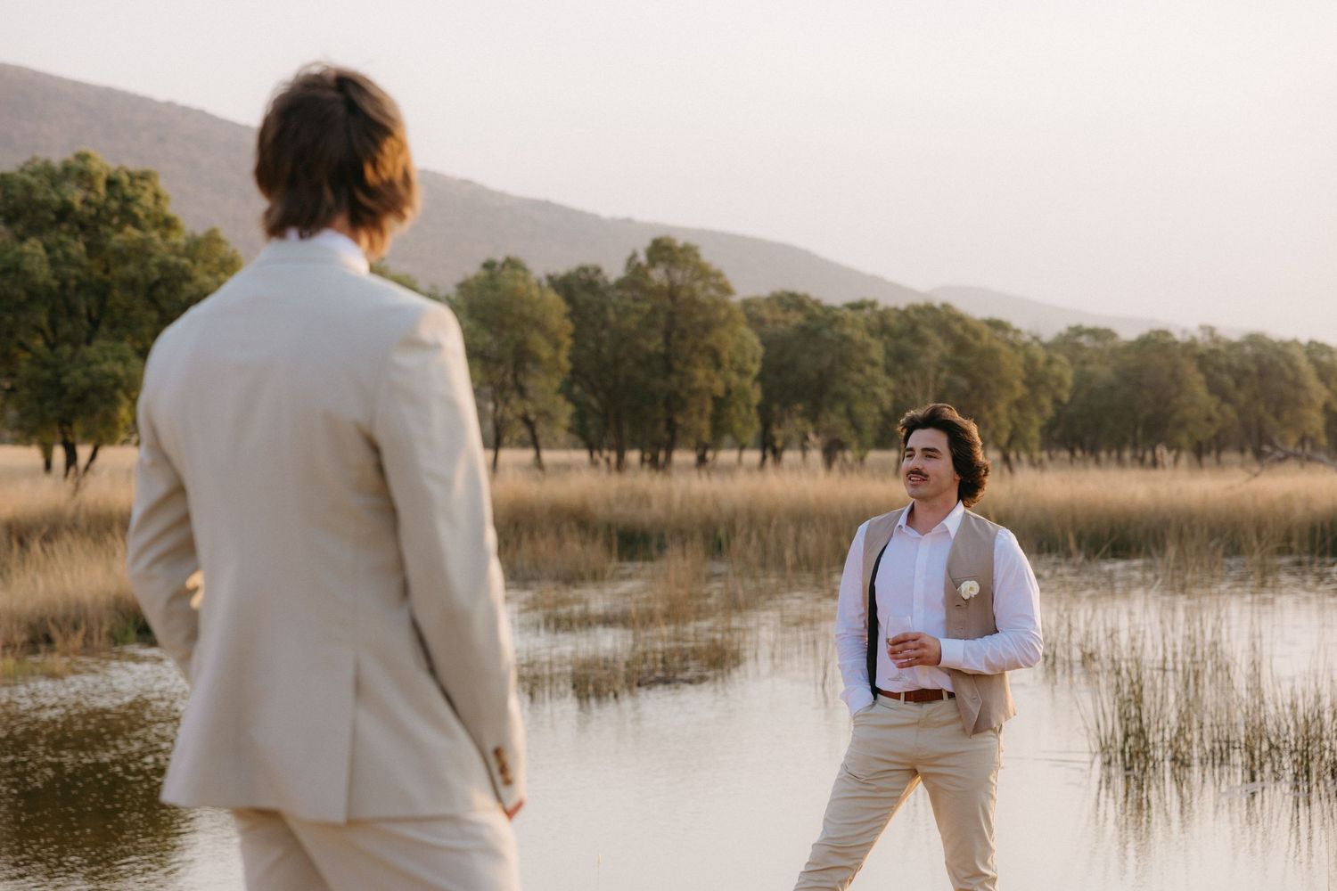 Couple in light colored attire walking along a serene lake shore at sunset in a natural outdoor setting.