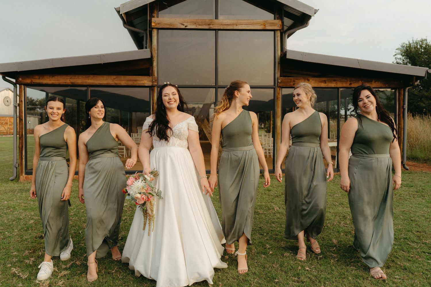 Bride and bridesmaids in sage green dresses posing together outside rustic venue.