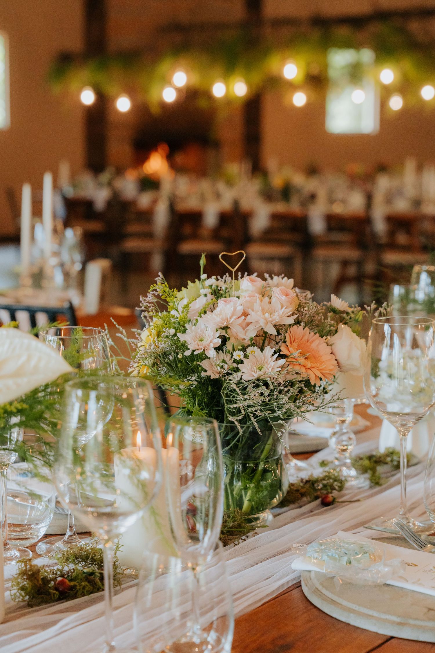 Elegant wedding reception table with blush floral centerpieces, white candles and string lights creating a romantic atmosphere.