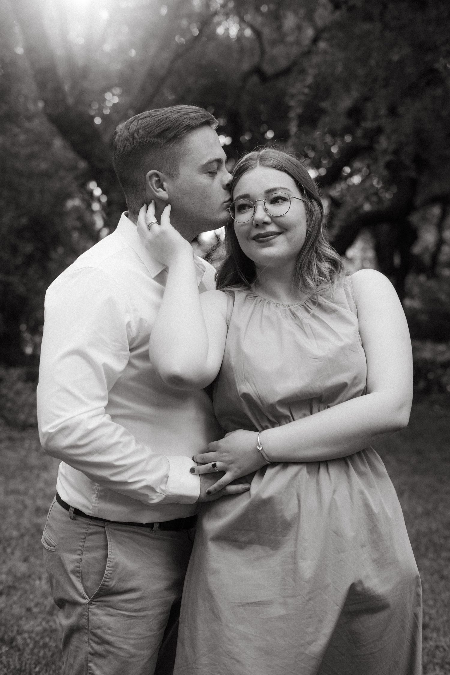 A romantic black and white portrait of a couple embracing outdoors in nature with sunlight filtering through trees.