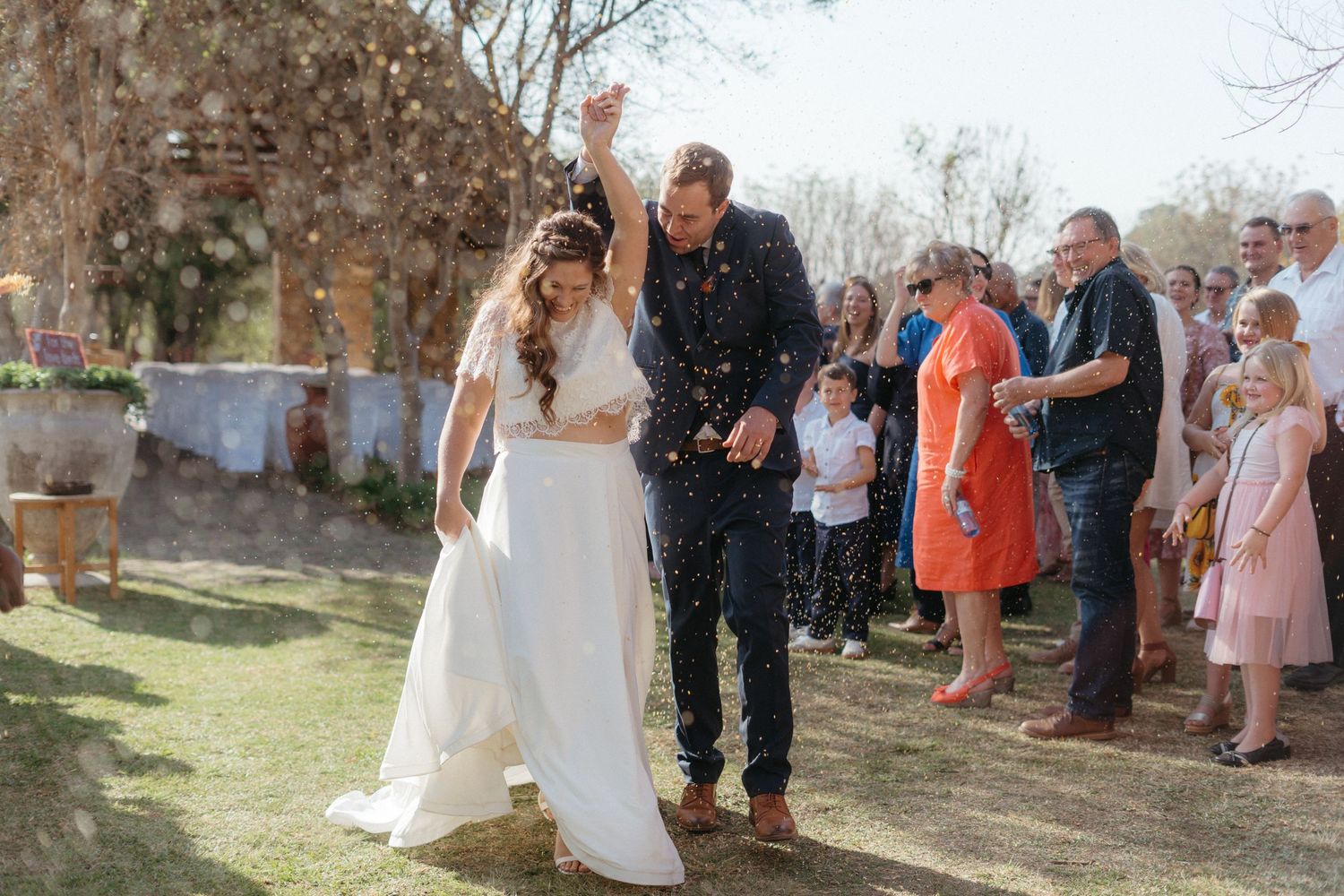 Newlyweds walking through shower of confetti with wedding guests in background.
