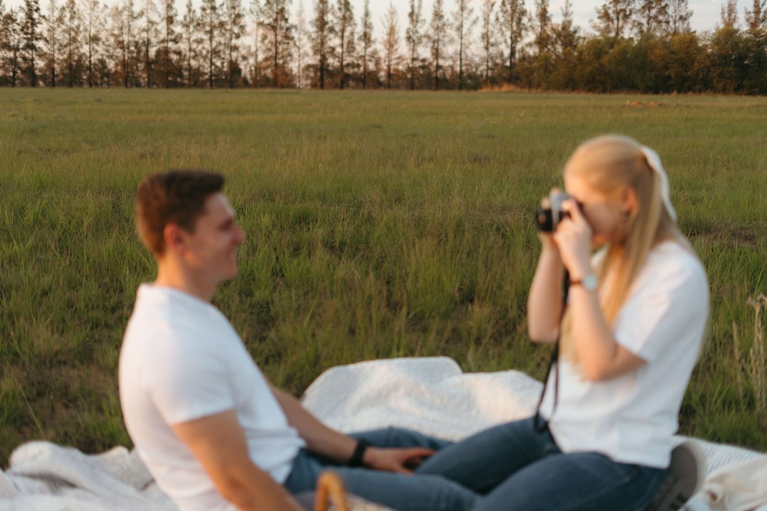 Two people sit on a white blanket in a grassy field during sunset with tall trees in the background.