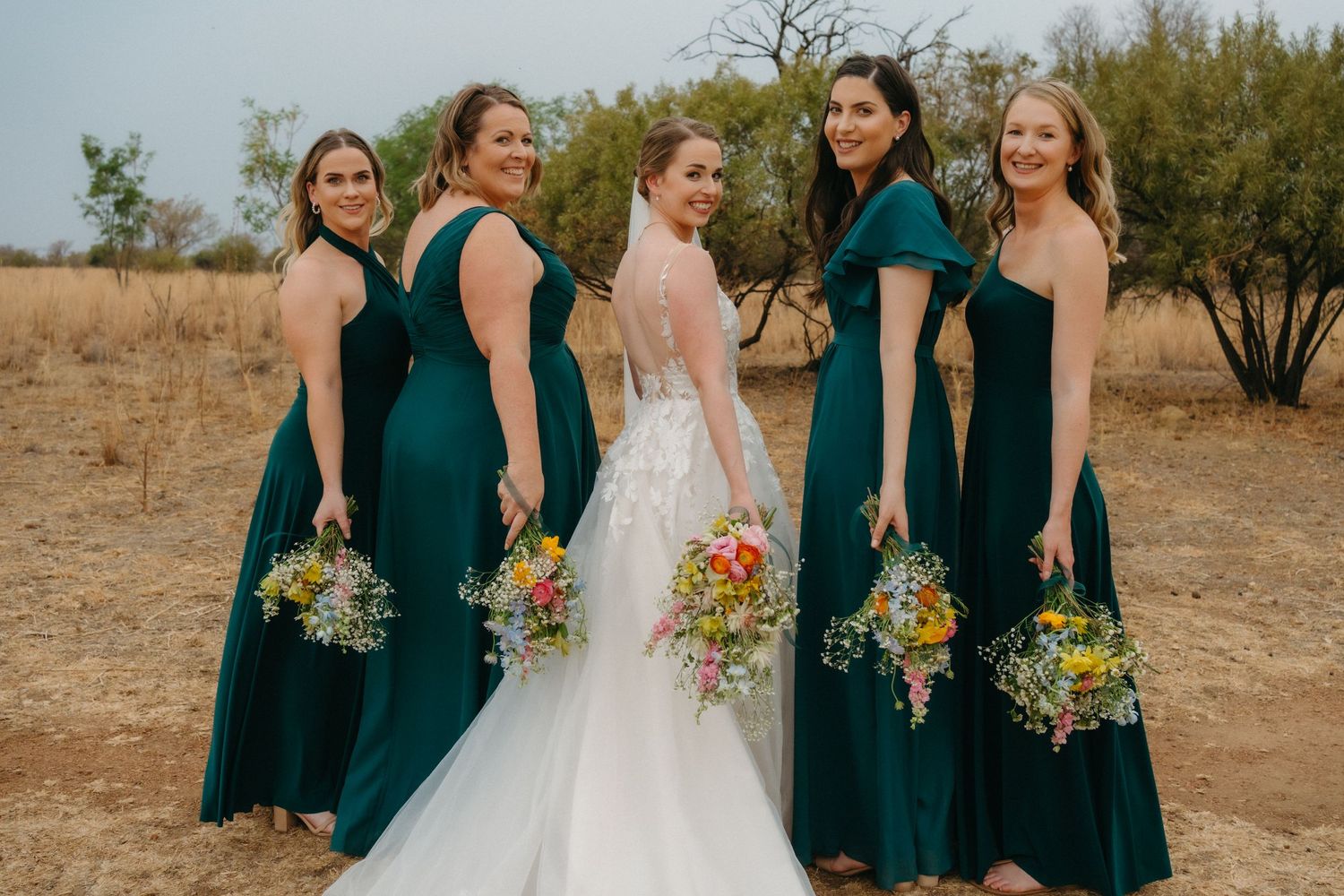 Bridesmaids in elegant emerald gowns hold vibrant bouquets while posing outdoors.