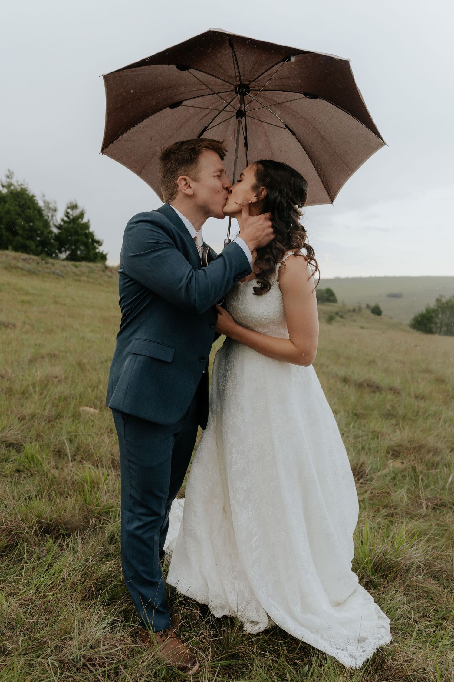 Newlywed couple shares romantic kiss under brown umbrella on grassy hillside during overcast wedding day.
