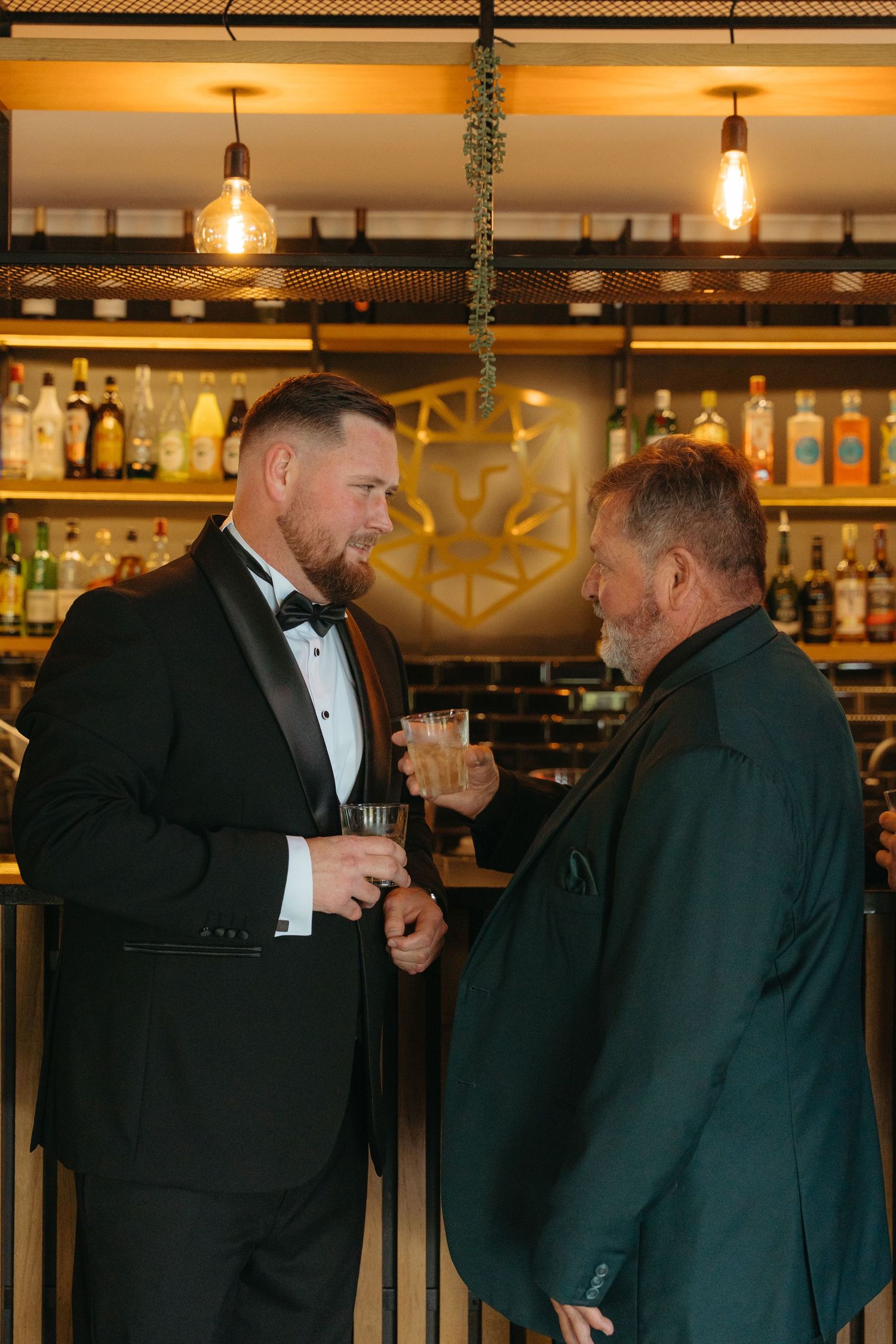 Two men in formal attire having a conversation at an elegant bar with warm lighting and bottles in the background.