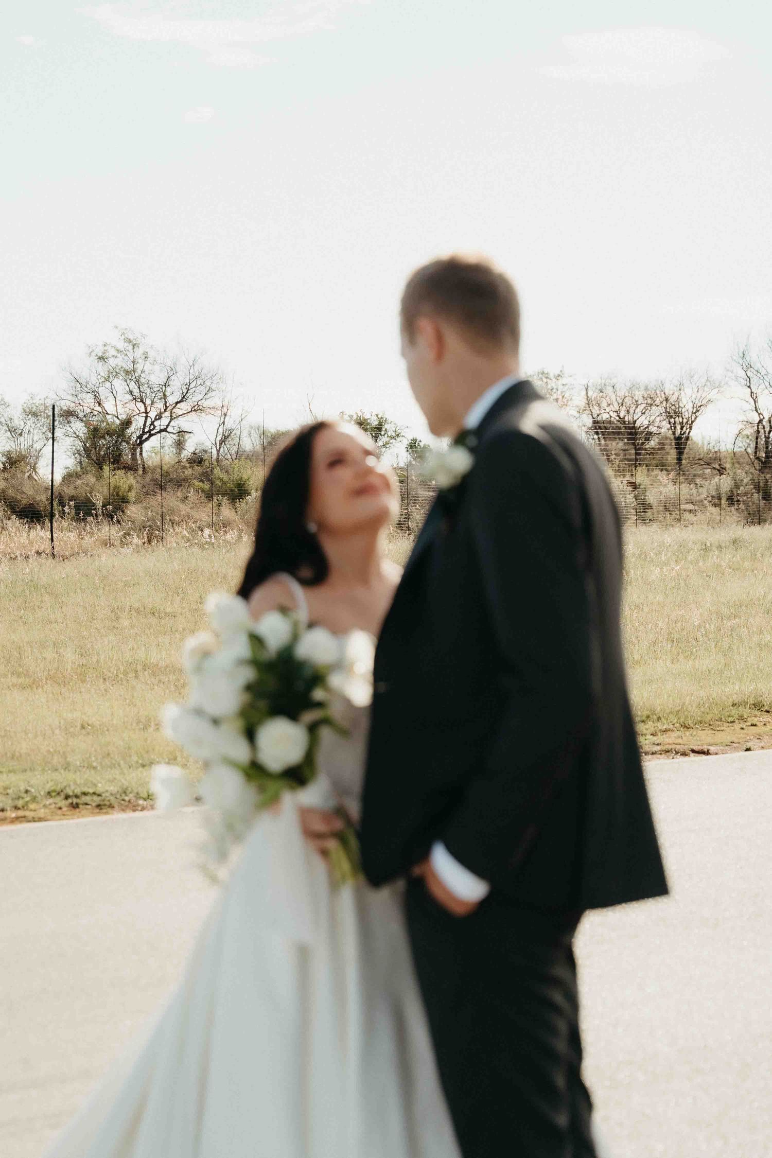 A bride in a white gown holds a bouquet of white roses while gazing at her groom in a black tuxedo outdoors.