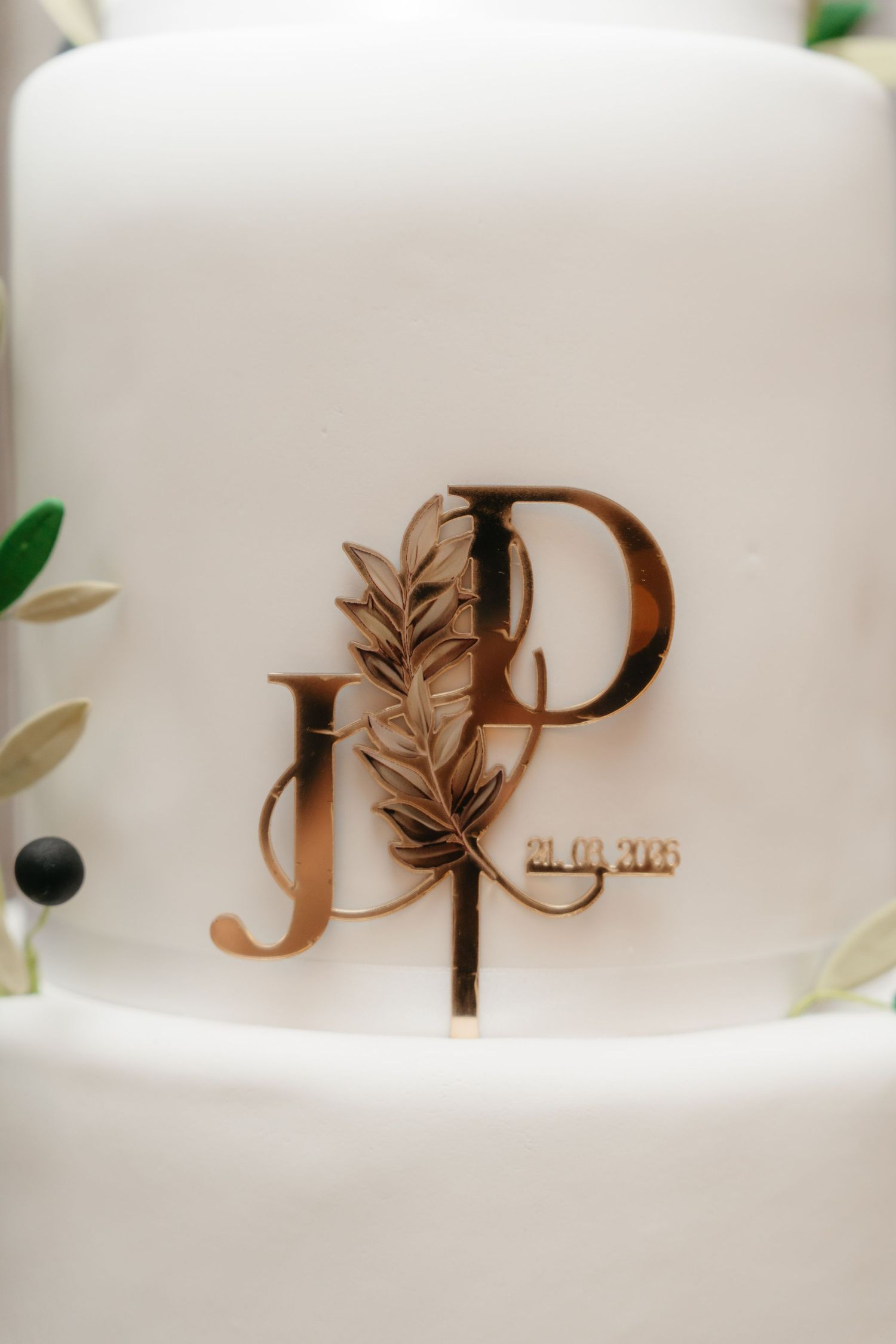 Gold monogram wedding cake topper with initials JP and date on elegant white tiered cake with olive branch decor.