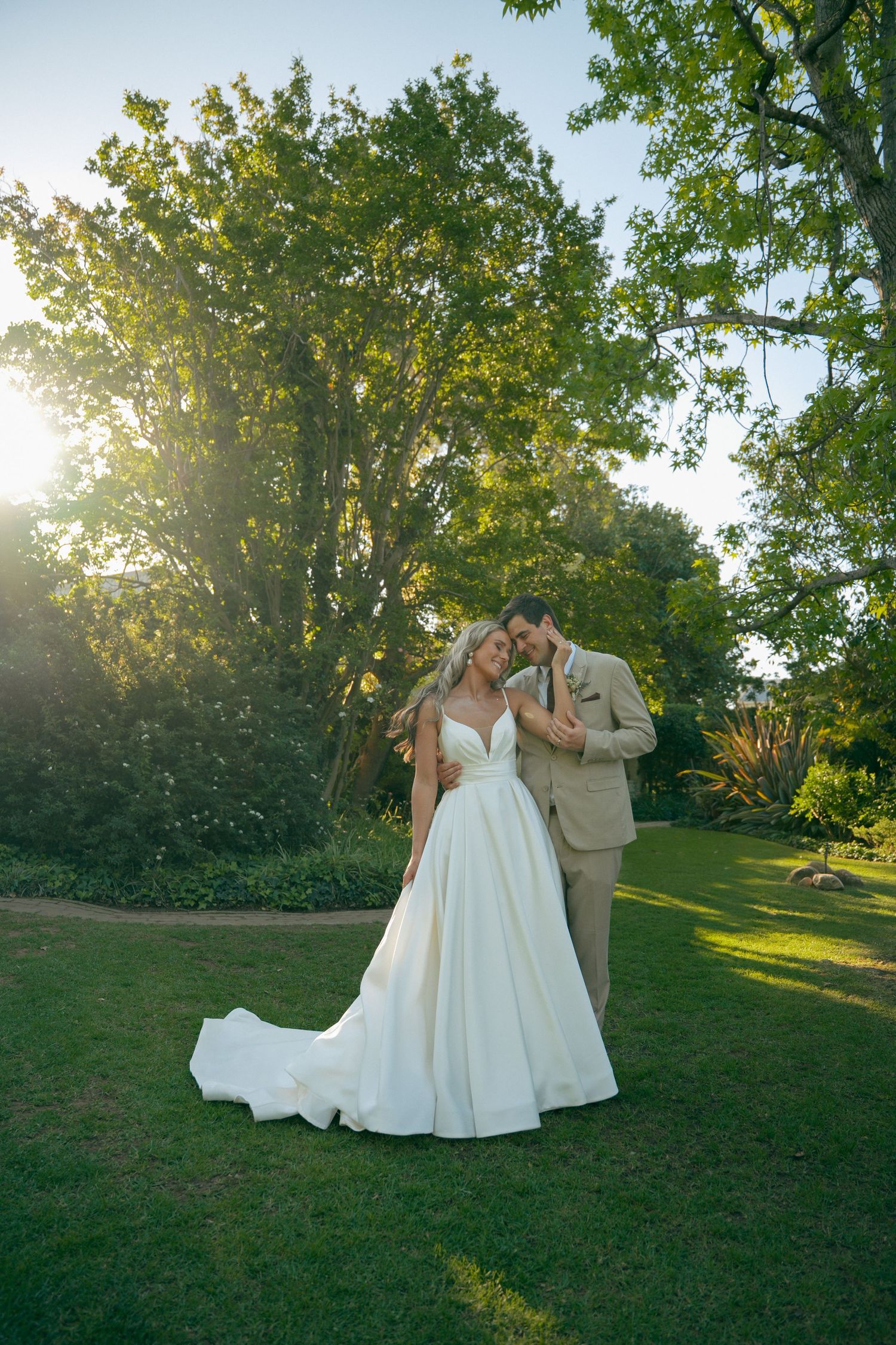 Bride in a white satin gown and groom in a tan suit embrace on a sunlit garden lawn surrounded by lush green trees.