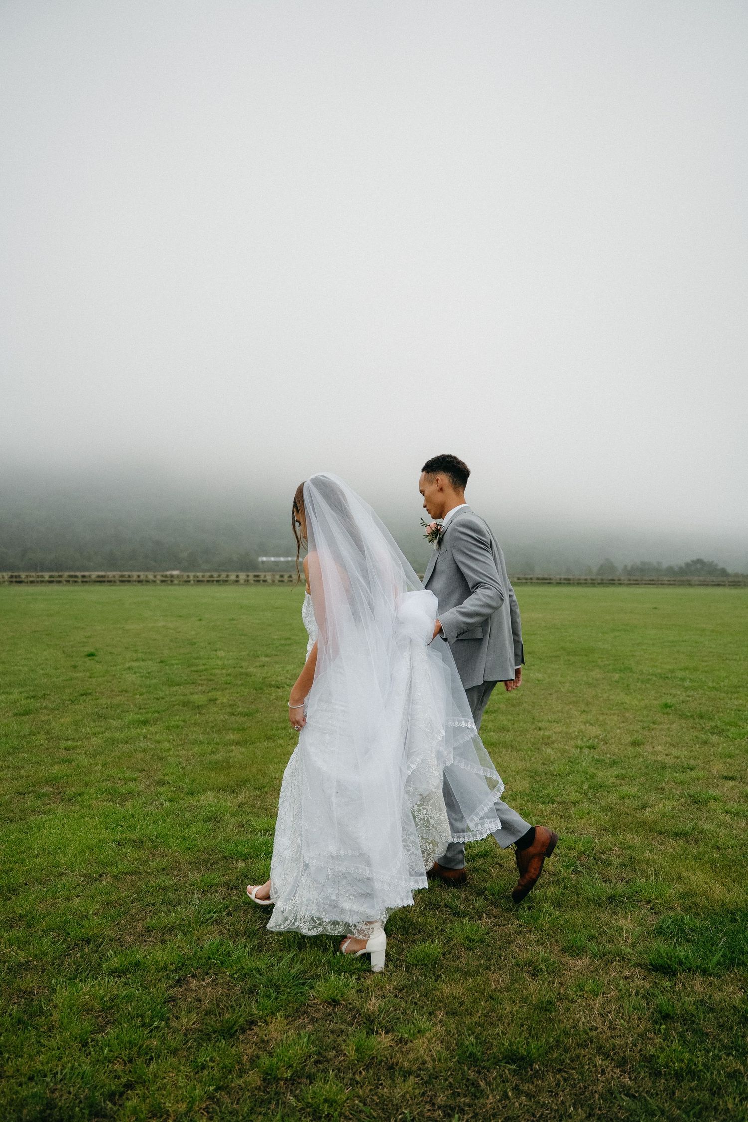 Bride in white lace gown and veil walks with groom in gray suit across misty green field.