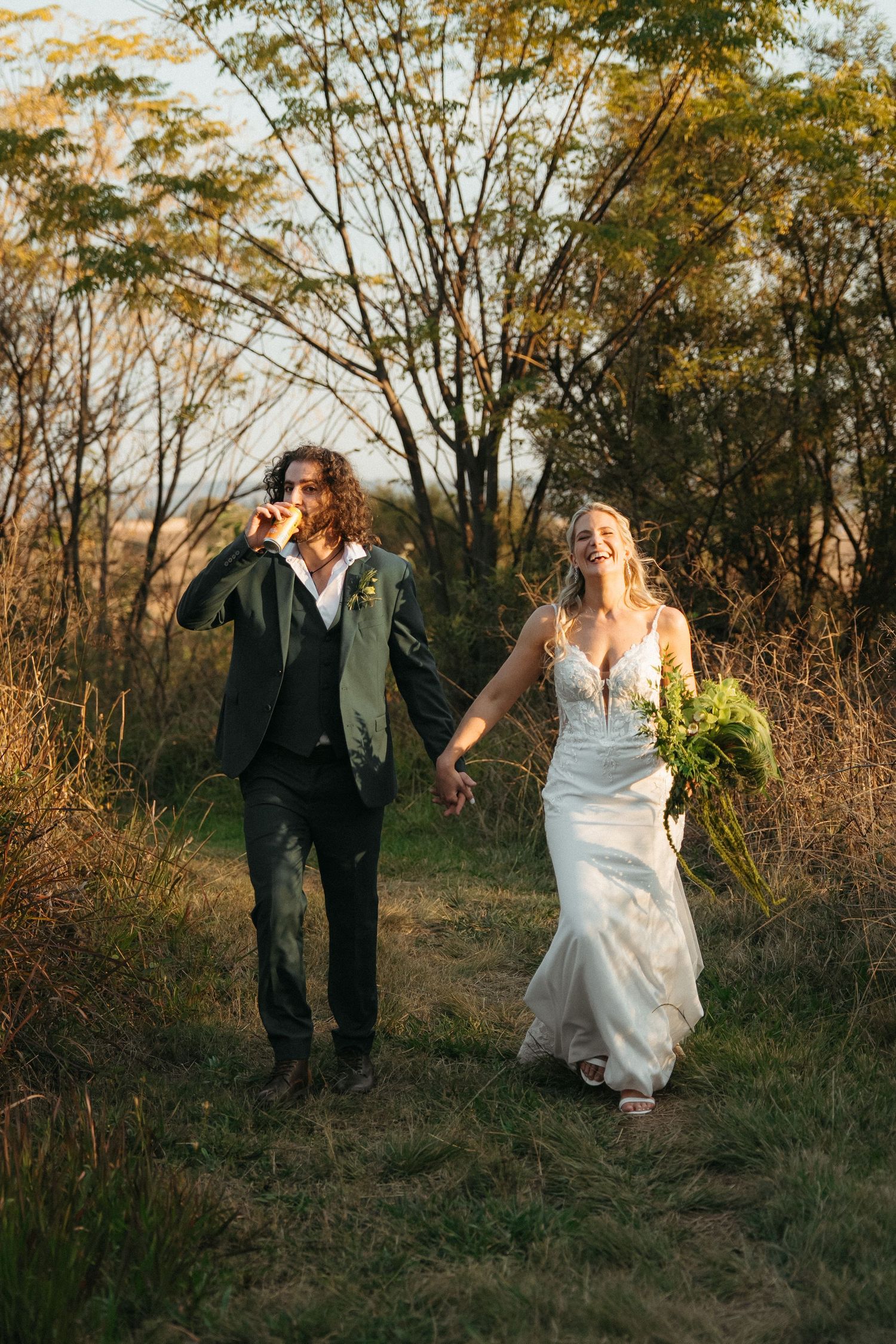 A laughing bride in a white gown and groom in a dark suit walk hand-in-hand through a golden meadow at sunset.