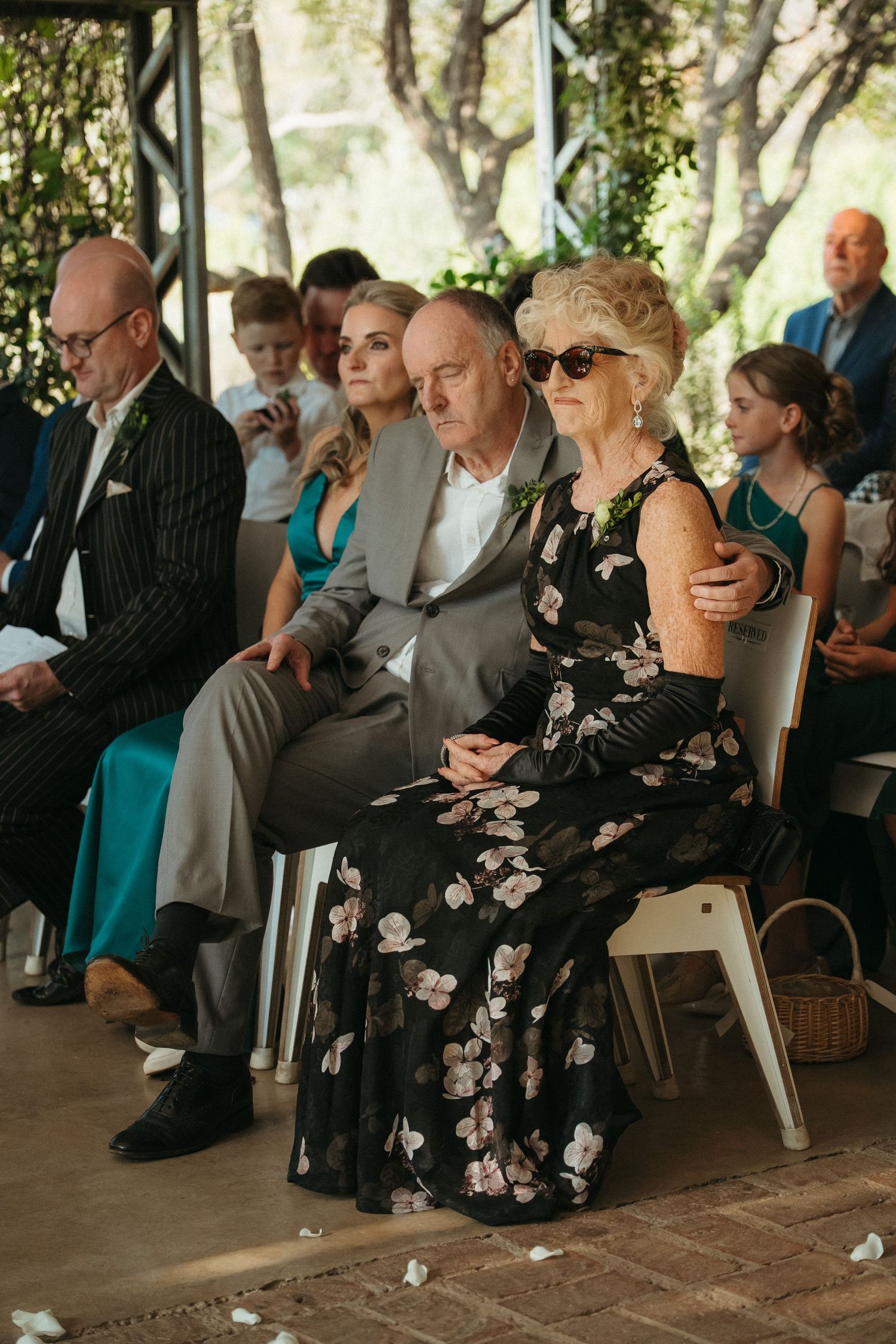 Elderly couple in formal attire seated at an outdoor wedding ceremony, woman in floral dress with sunglasses.