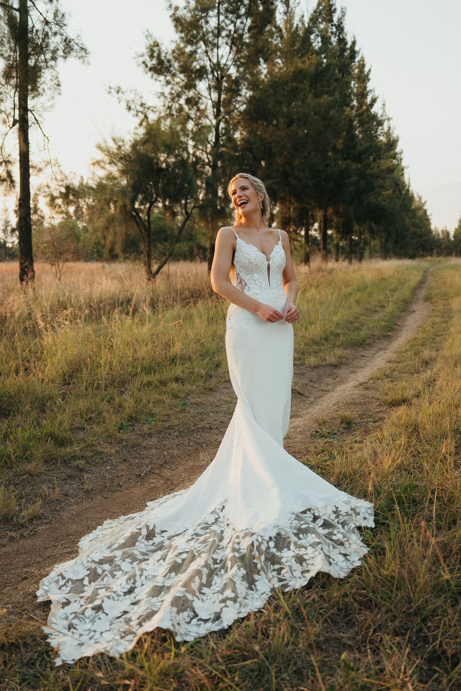 Laughing bride in a white lace mermaid wedding gown with a train stands on a dirt path surrounded by trees.