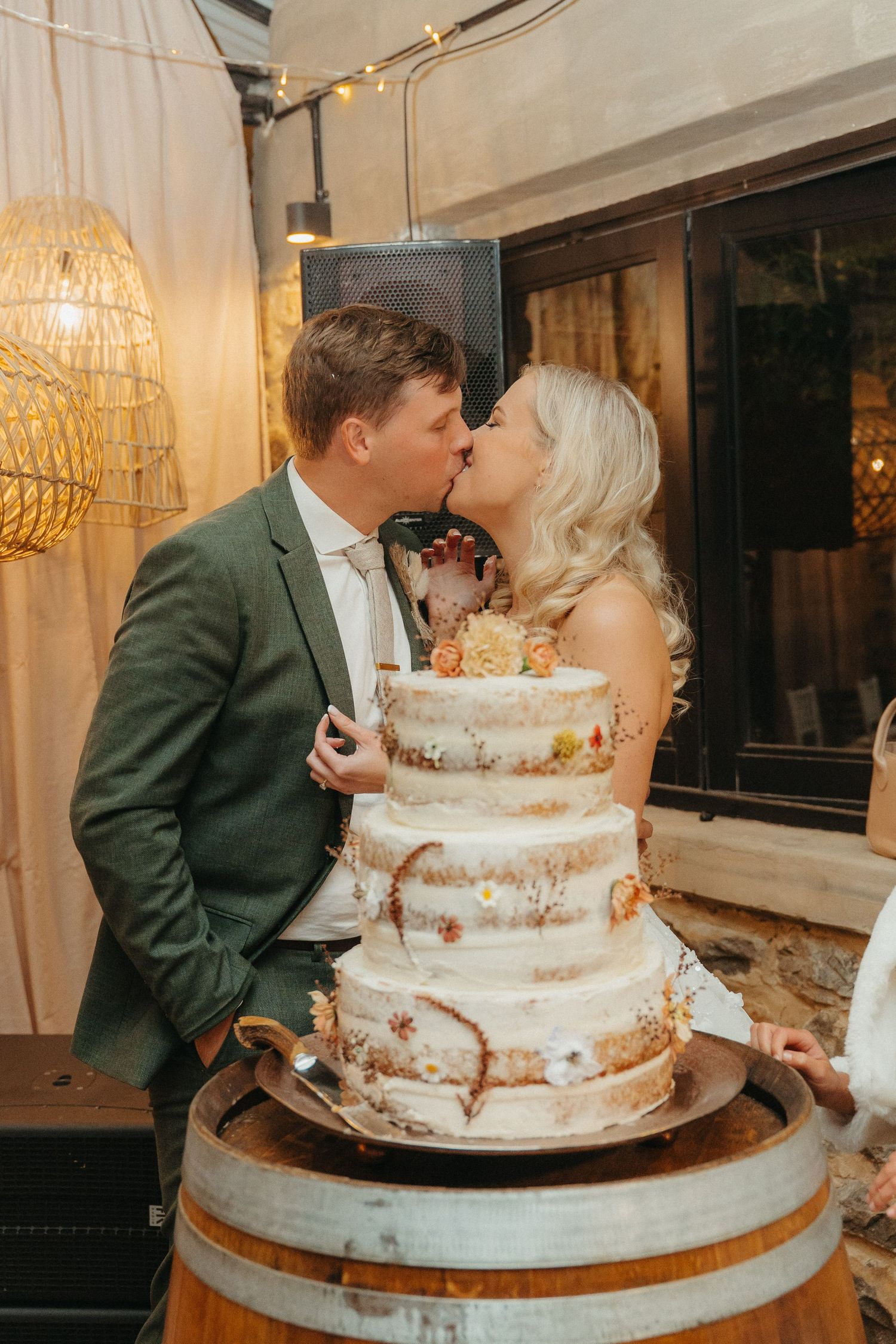 Newlyweds share a kiss behind a rustic three-tier semi-naked wedding cake adorned with flowers on a barrel.