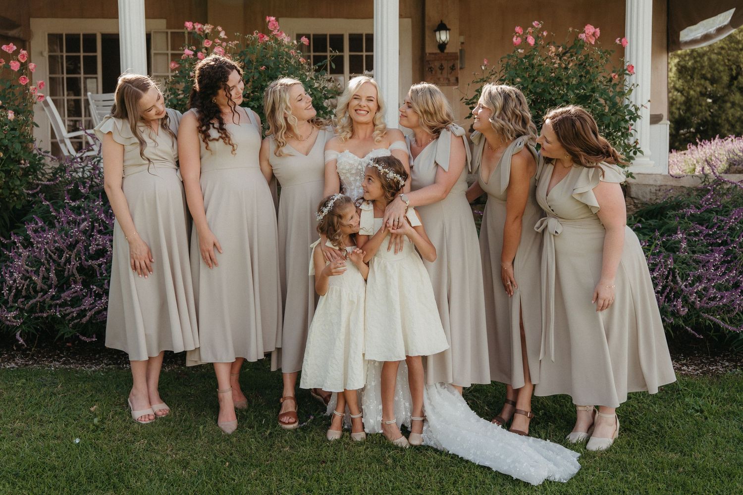 Bride with bridesmaids in sage green dresses and two flower girls in white dresses posing outdoors at a wedding.