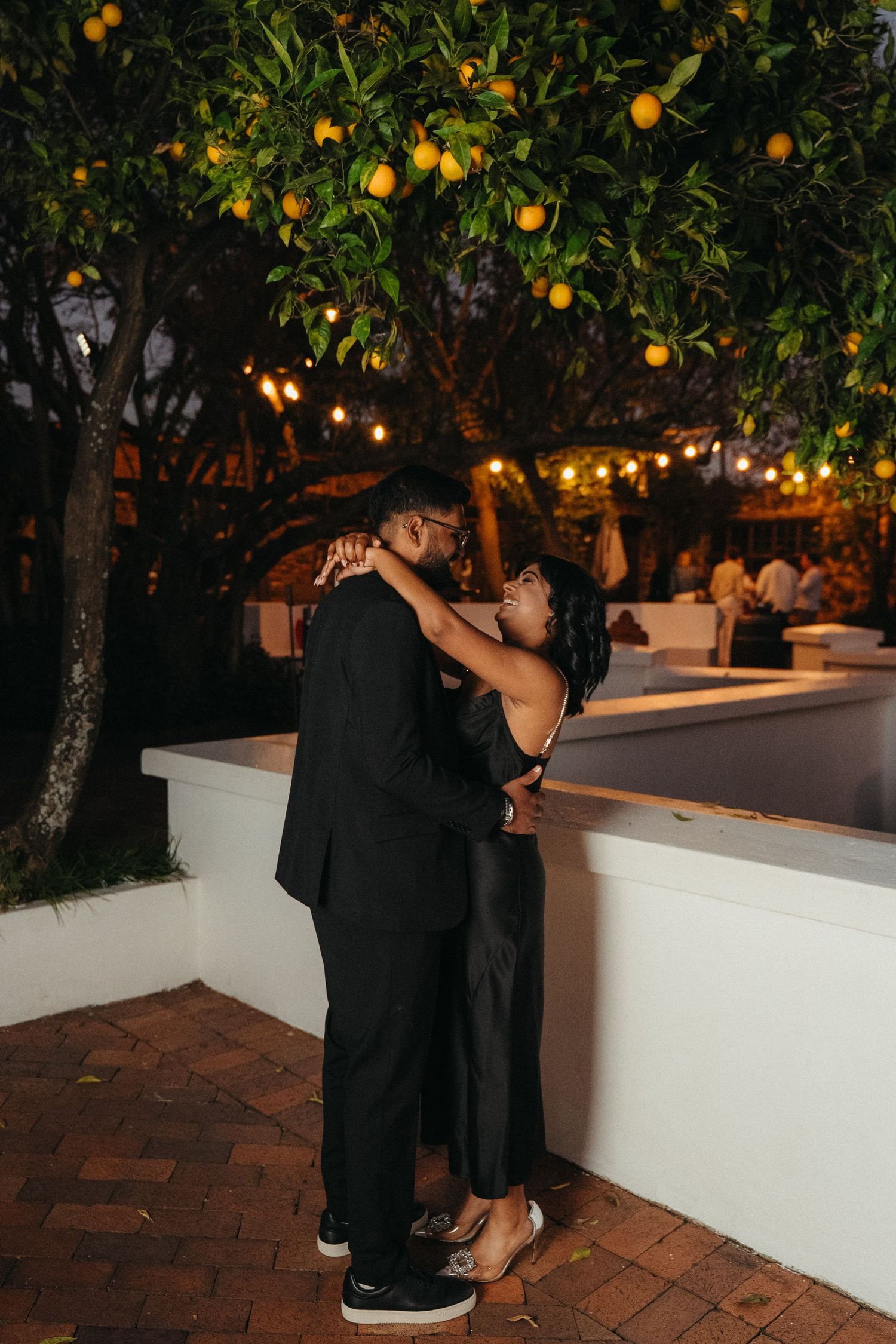 Couple embracing romantically under a lemon tree at night, dressed in black formal attire with string lights glowing.