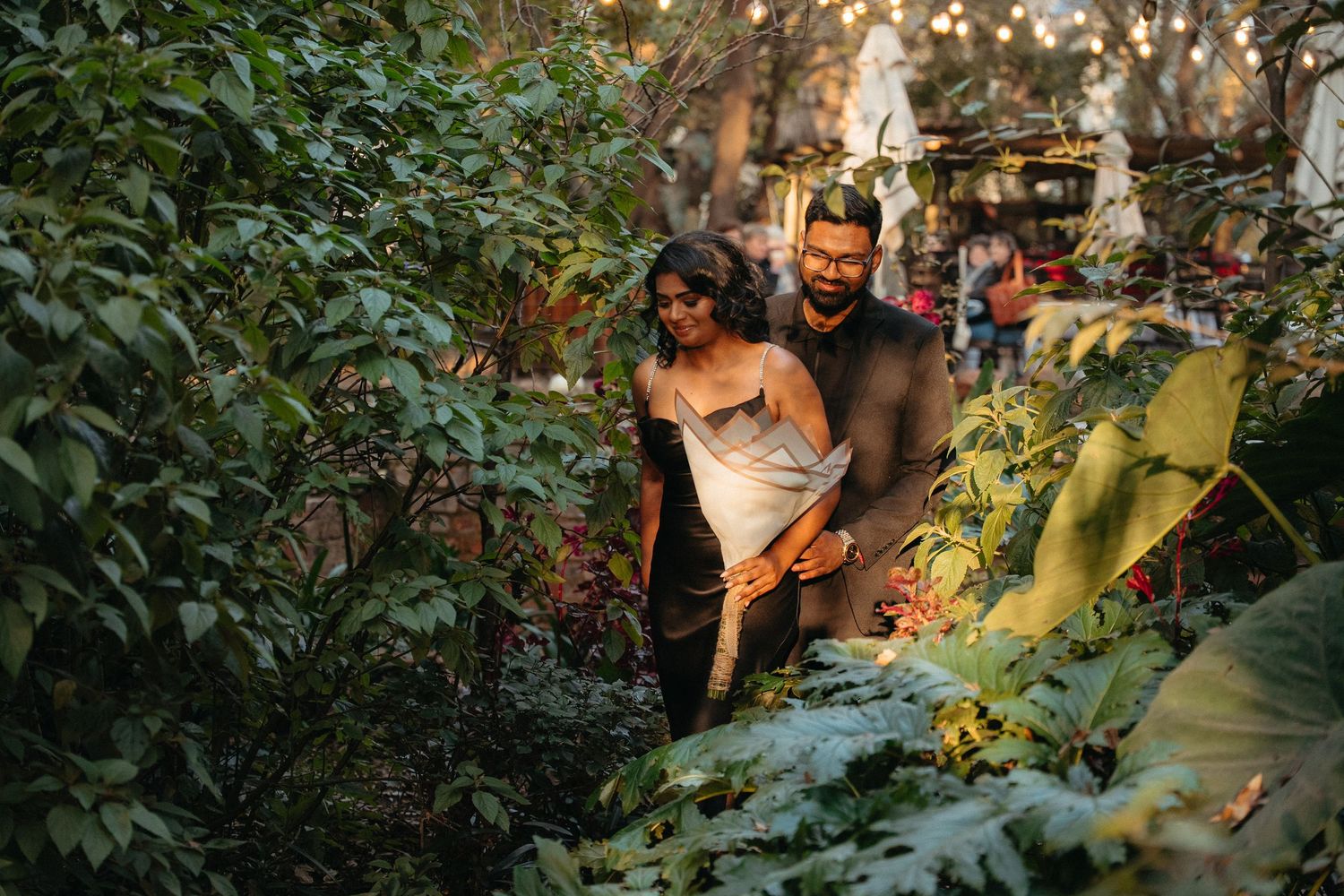 Couple walking through lush garden greenery, woman in white dress and man in casual attire, surrounded by tropical plants.