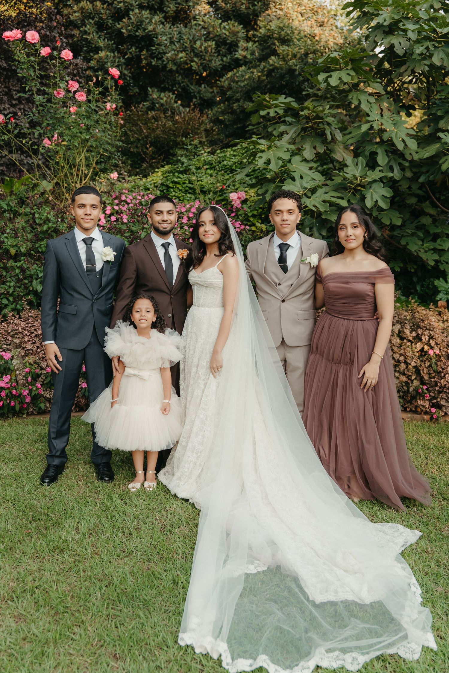 Wedding party poses in garden, bride in white gown with veil, groom in brown suit, groomsmen, bridesmaid, and flower girl.
