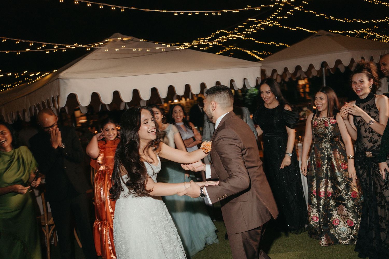 Bride and groom share a joyful first dance at an outdoor wedding reception under warm string lights.